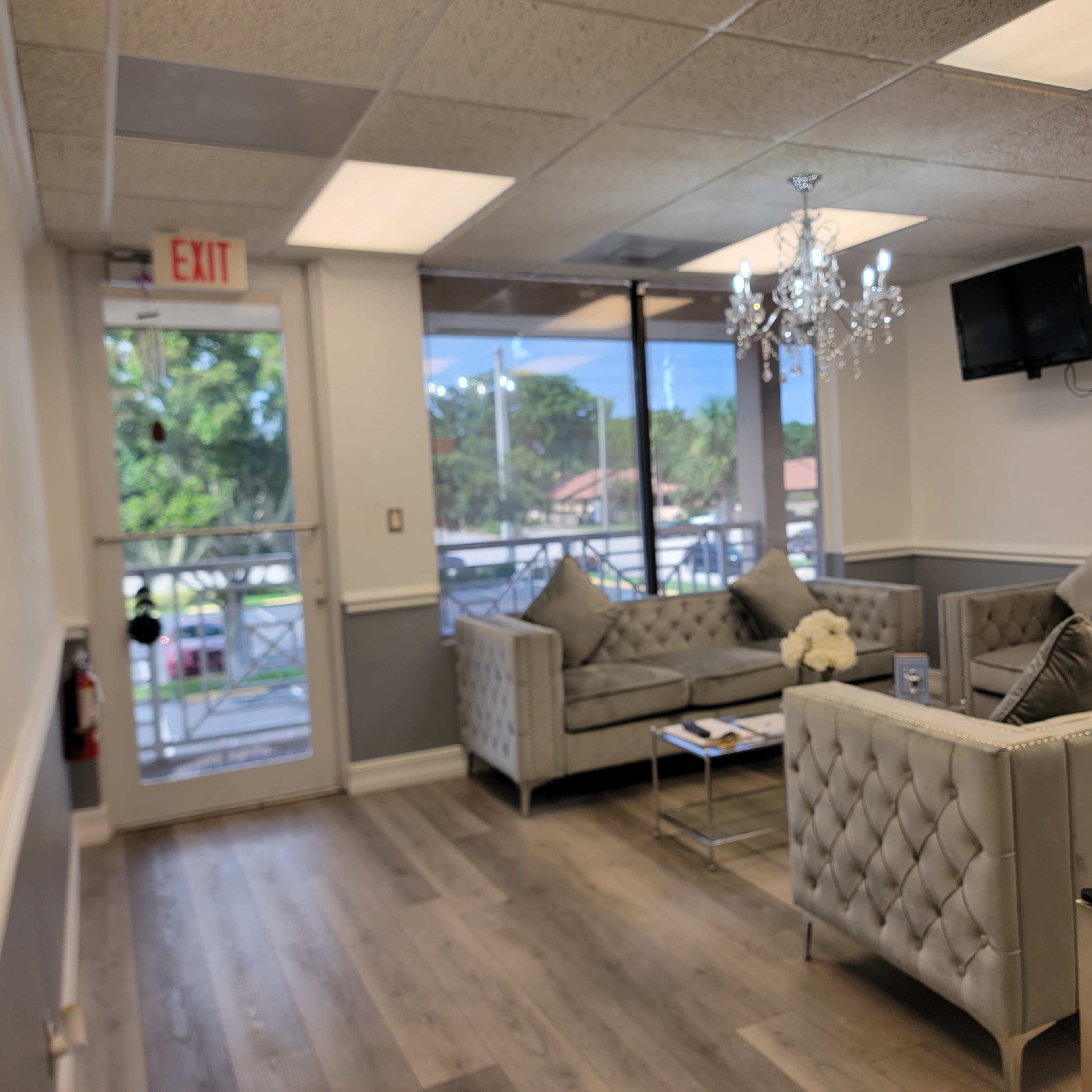 A waiting area with two gray tufted couches, a chandelier, a television, and large windows letting in natural light.