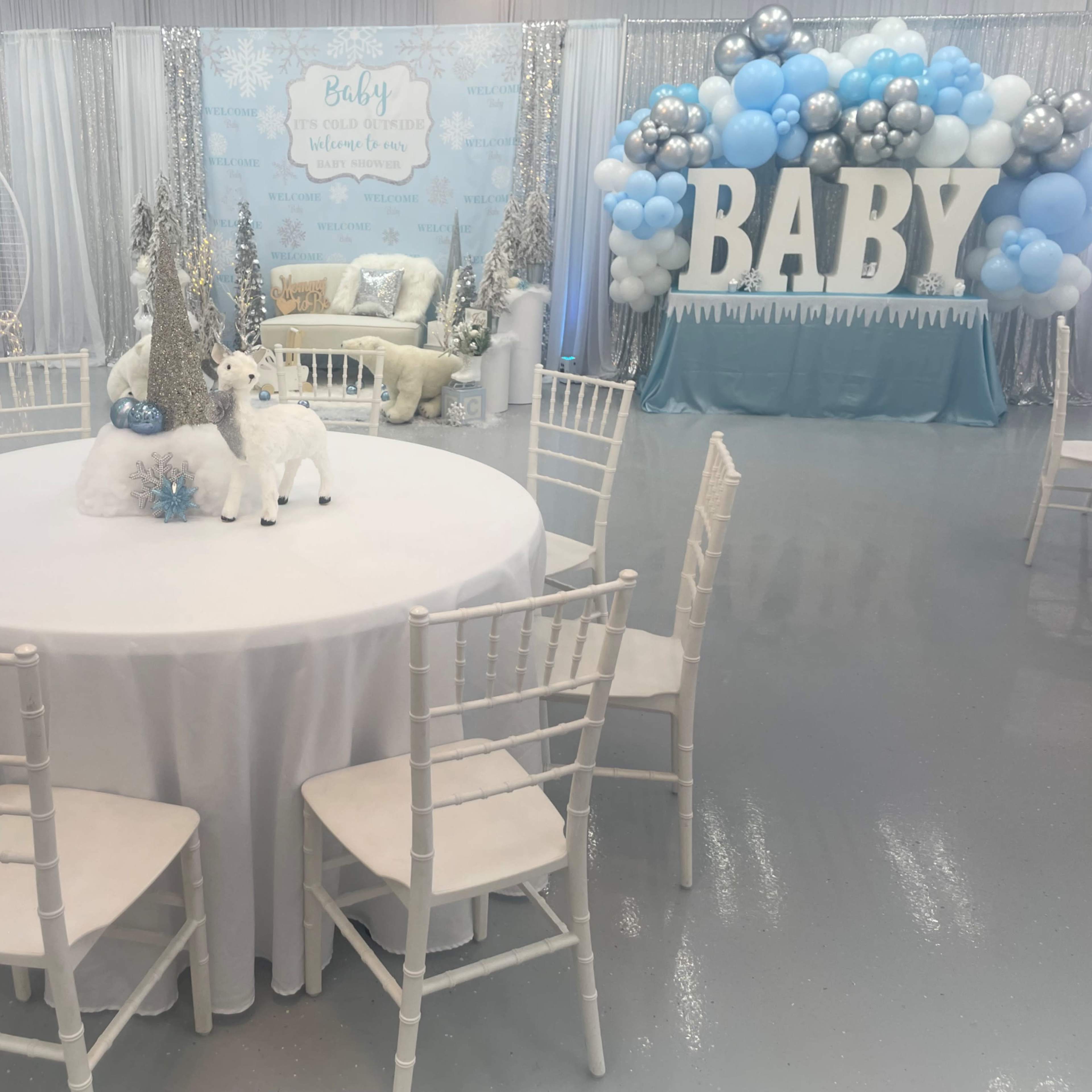 A decorated event space for a baby shower features a round table with a white tablecloth and centerpieces, surrounded by white chairs, and adorned with snow-themed decorations and a balloon arch displaying the word "BABY."