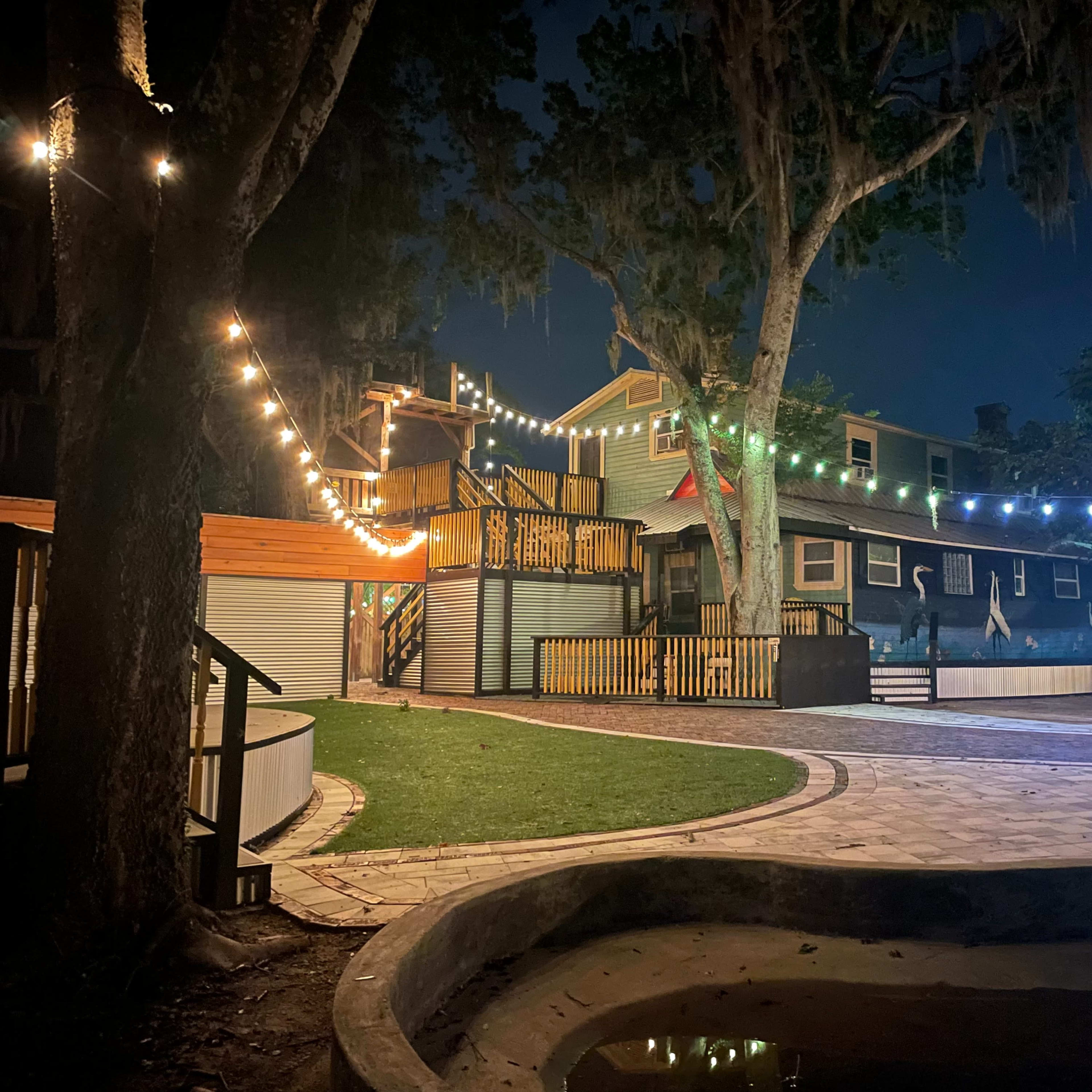 The scene shows a landscaped backyard at night, featuring a house illuminated by string lights and a circular patio area with grass and a small pond.