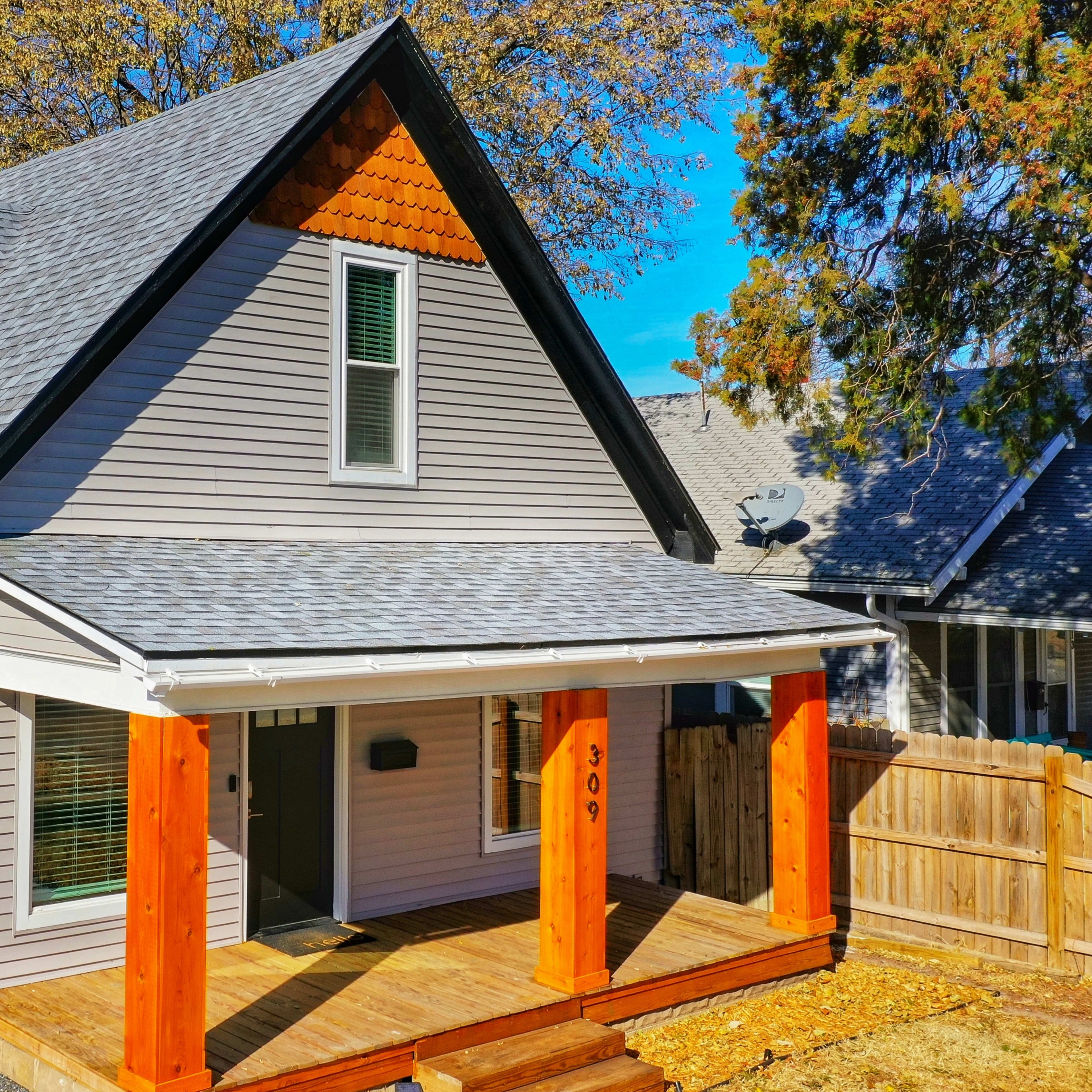 A two-story gray house with a peaked roof and wooden porch is set against a clear blue sky.