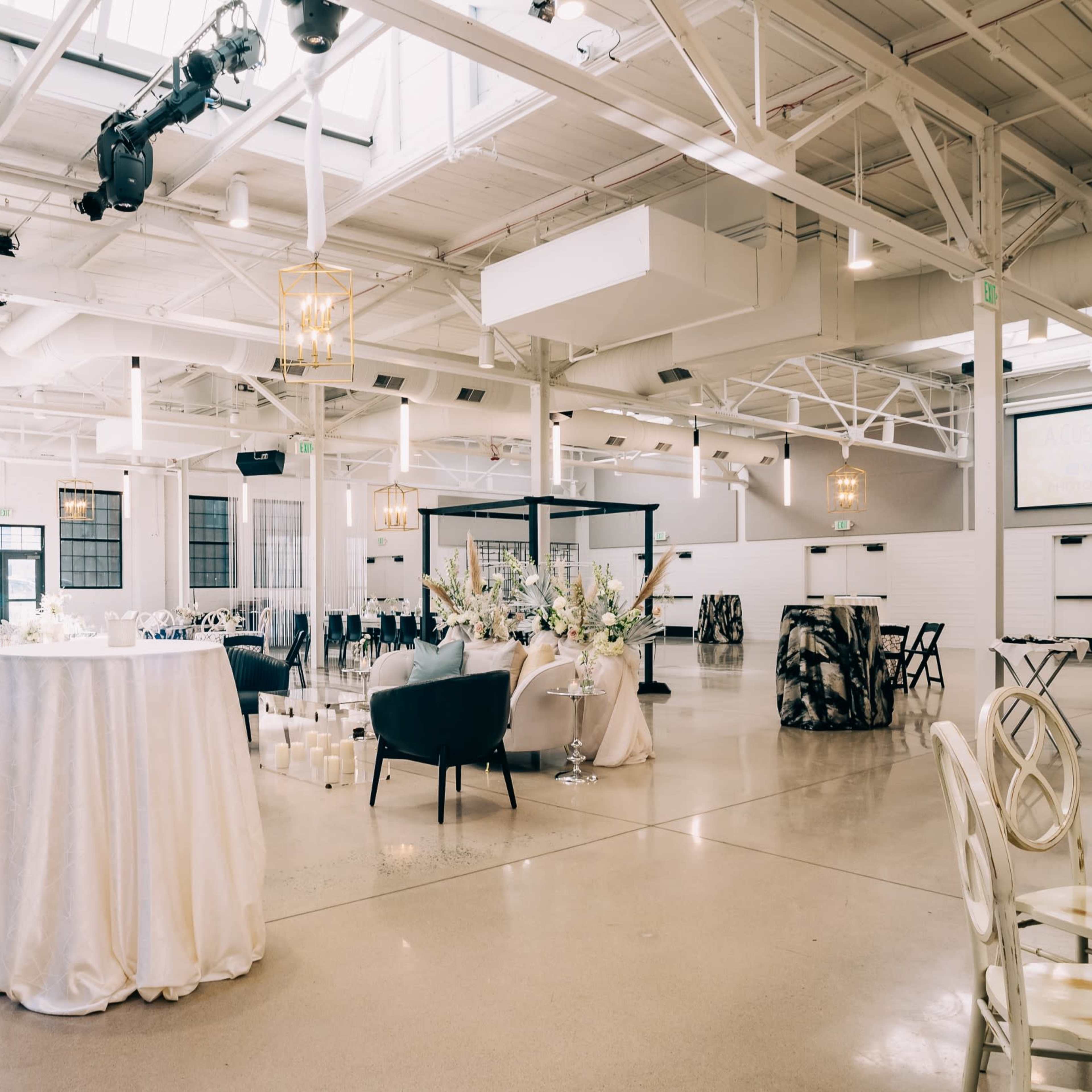A spacious event venue featuring elegantly arranged tables, chairs, and decorative elements under a bright, airy atmosphere created by large windows.