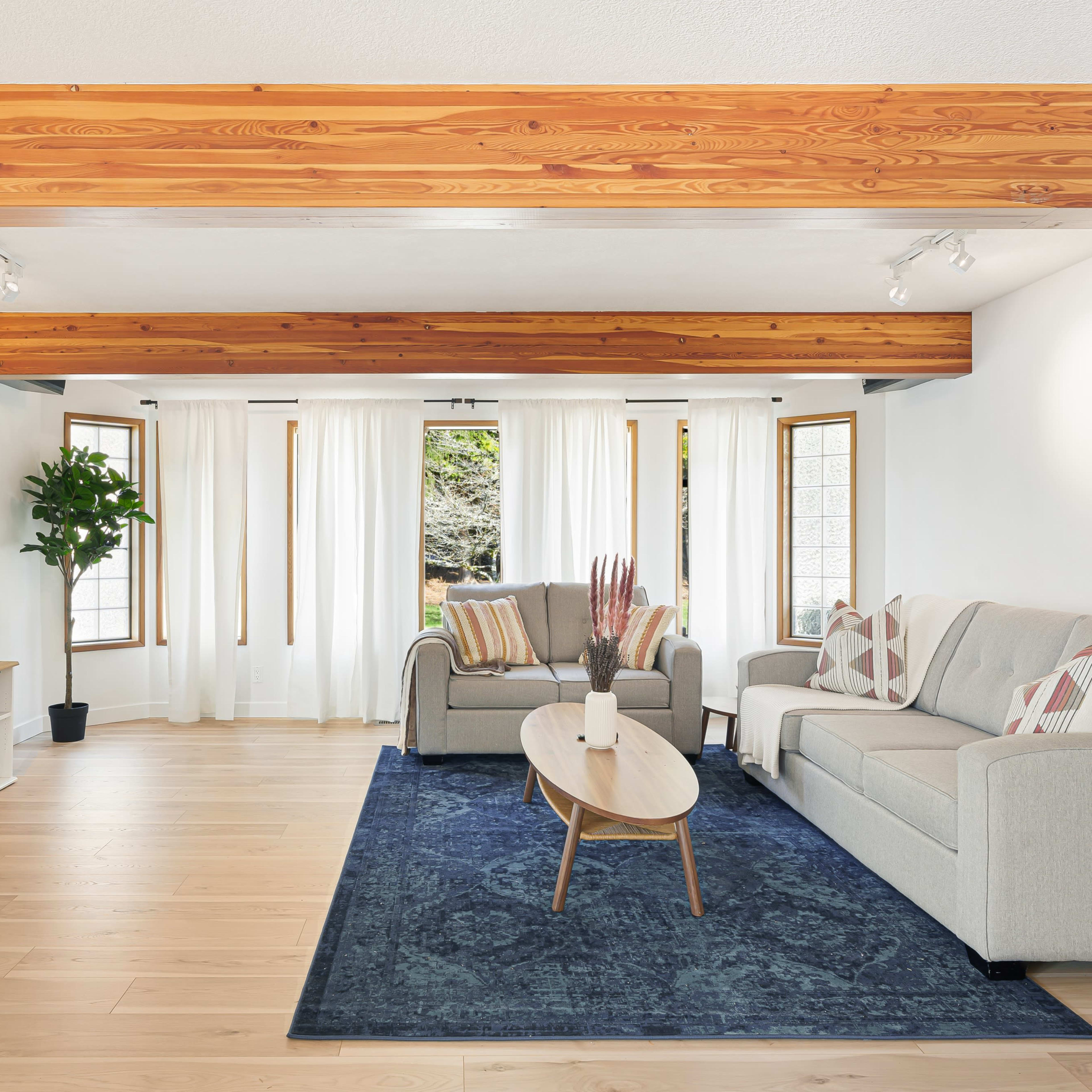 The room features a modern living area with a large television mounted on the wall, a sectional sofa, a coffee table, and large windows allowing natural light to flow in.