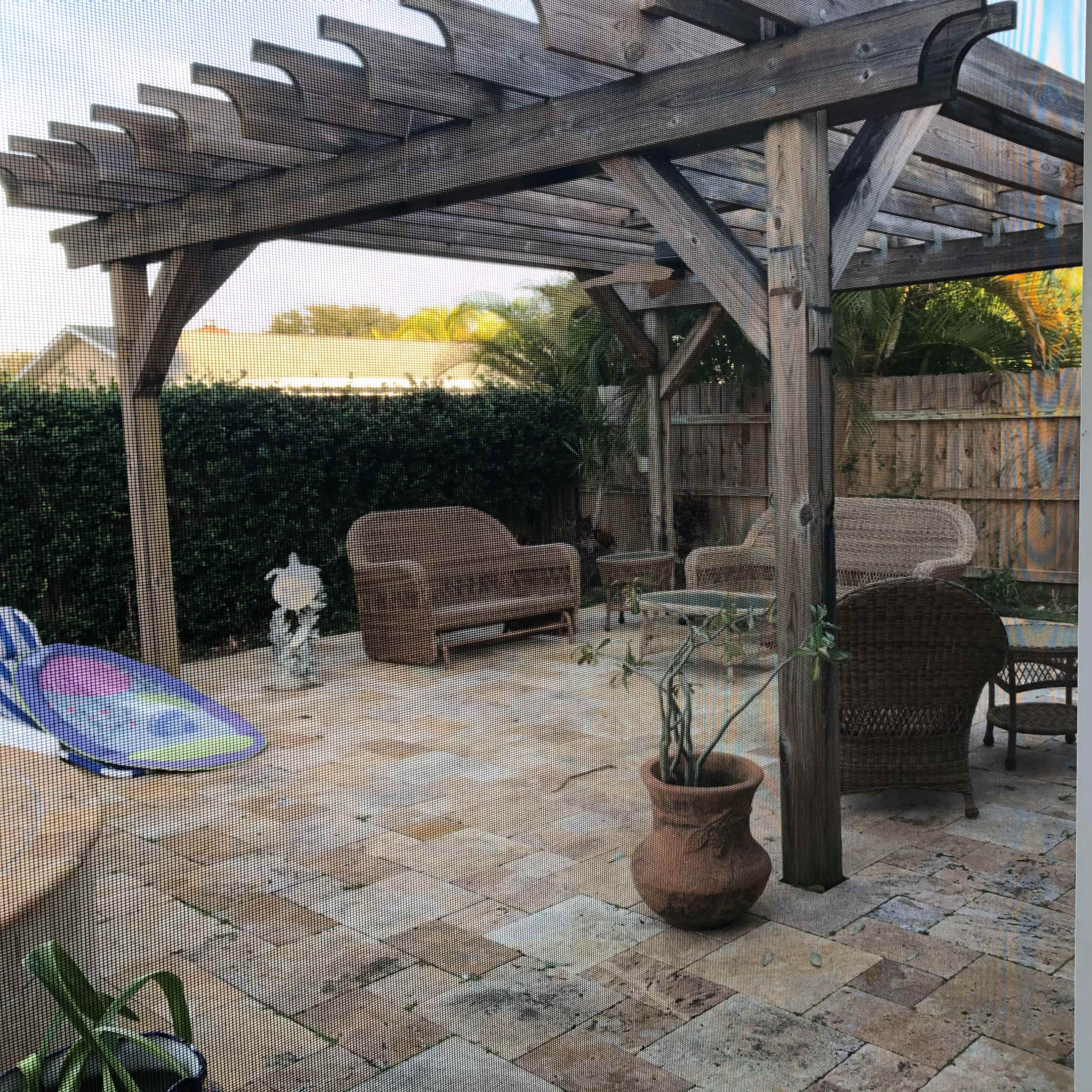 A patio area with a wooden pergola, comfortable seating, and potted plants, surrounded by hedges and a stone floor.