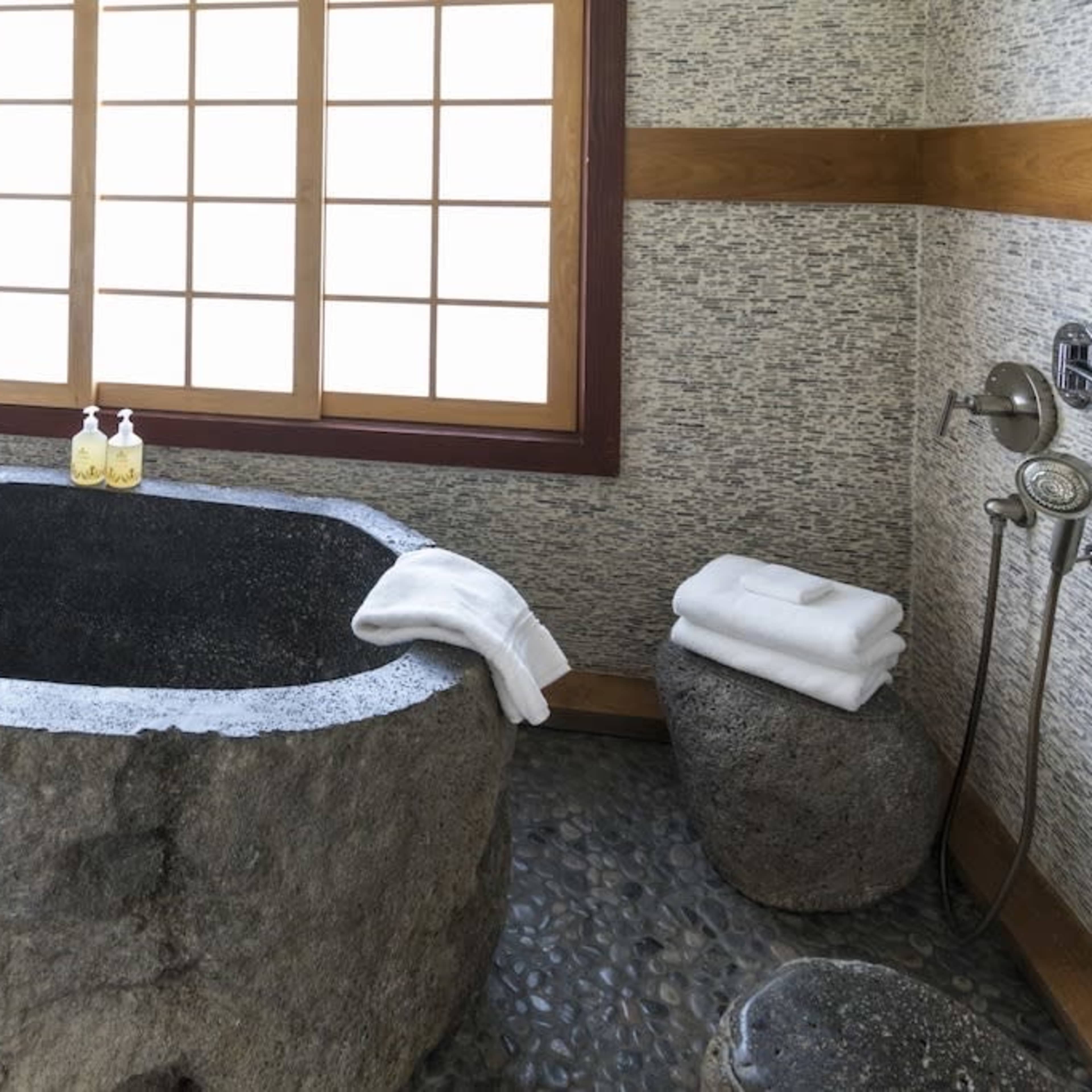 A minimalist bathroom featuring a stone bathtub, a shower with a chrome fixture, and neatly stacked white towels.