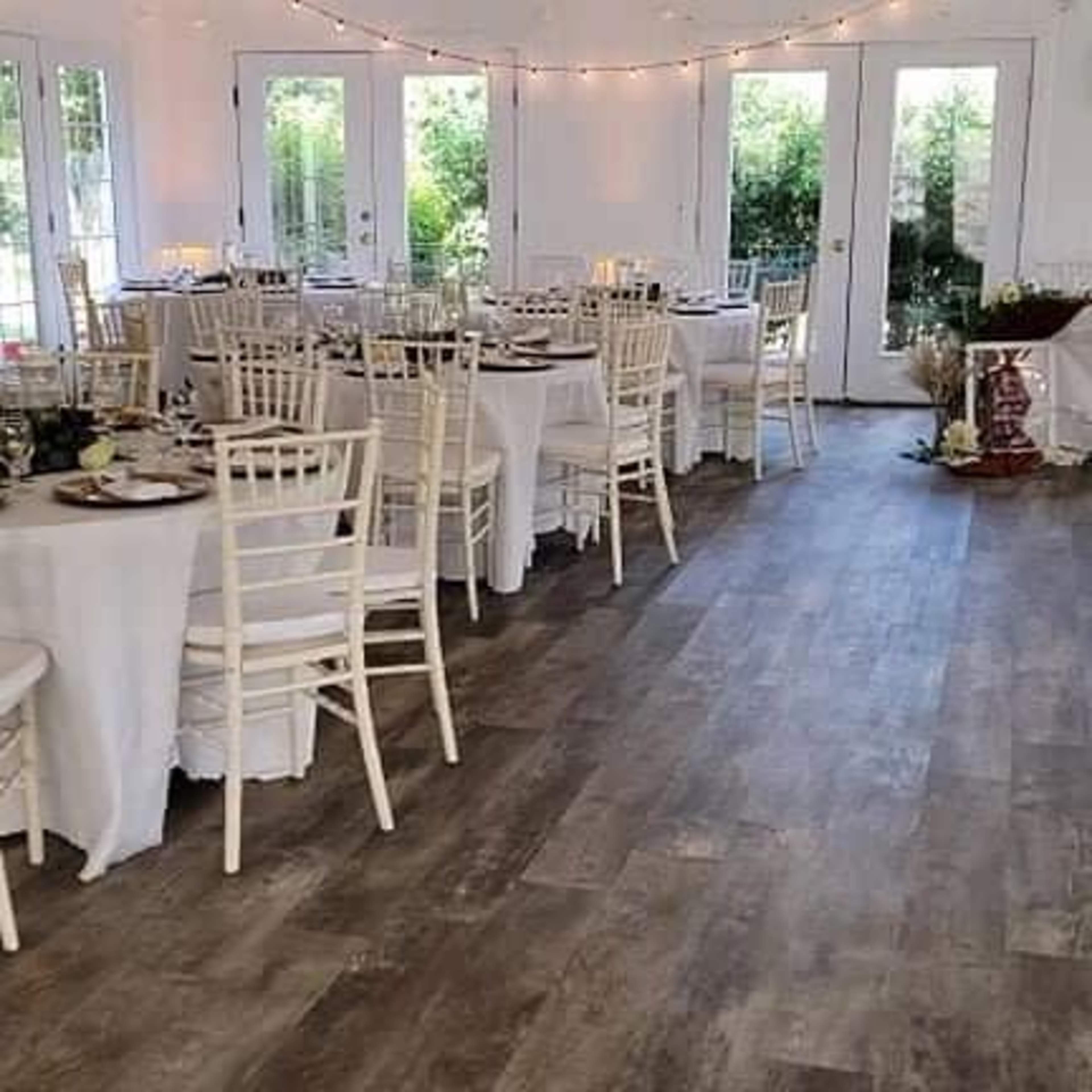 A banquet hall set up with round tables covered in white tablecloths, surrounded by wooden chairs, and decorated with string lights and greenery.