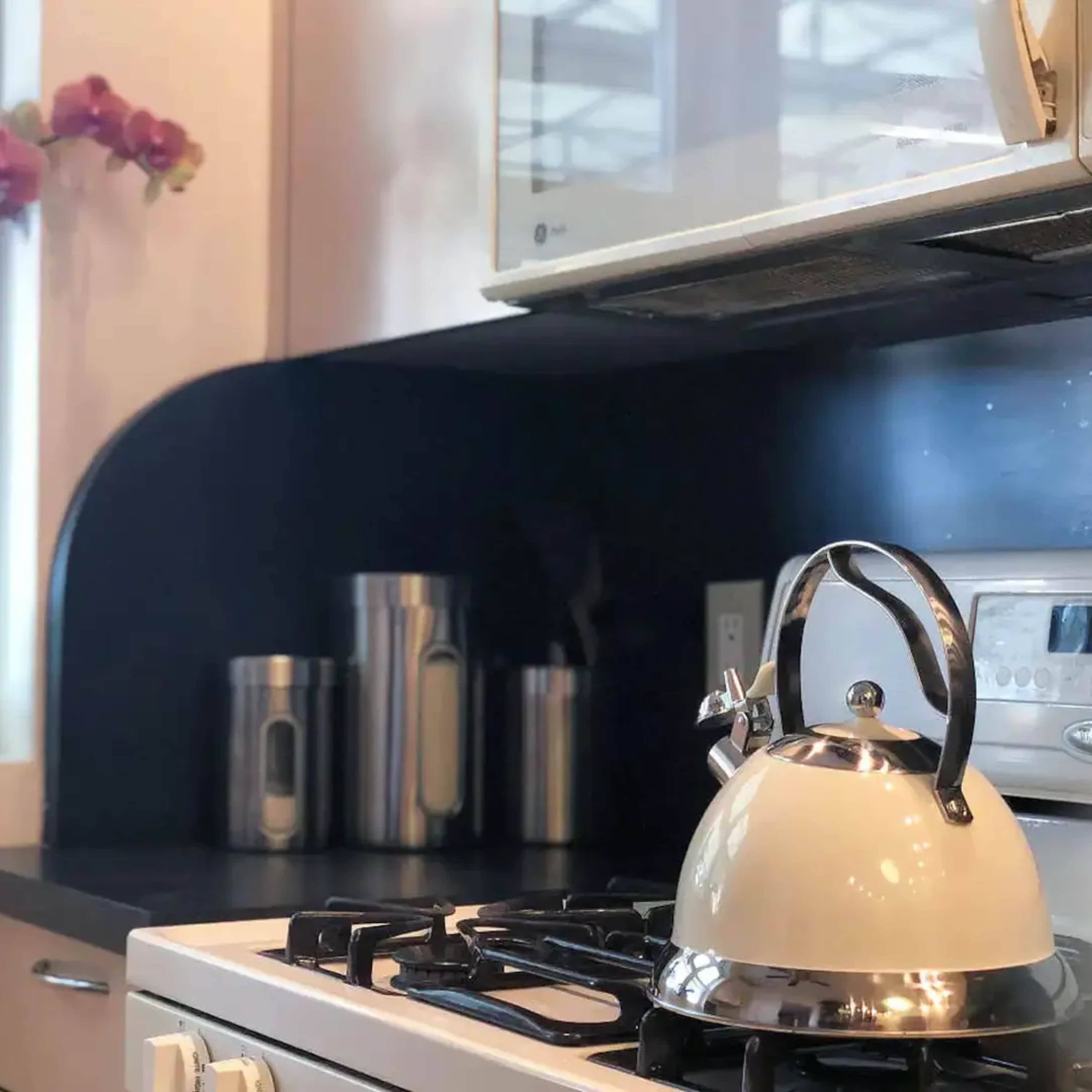 A kettle sits on a stovetop next to a collection of metal containers and a potted orchid in a brightly lit kitchen space.