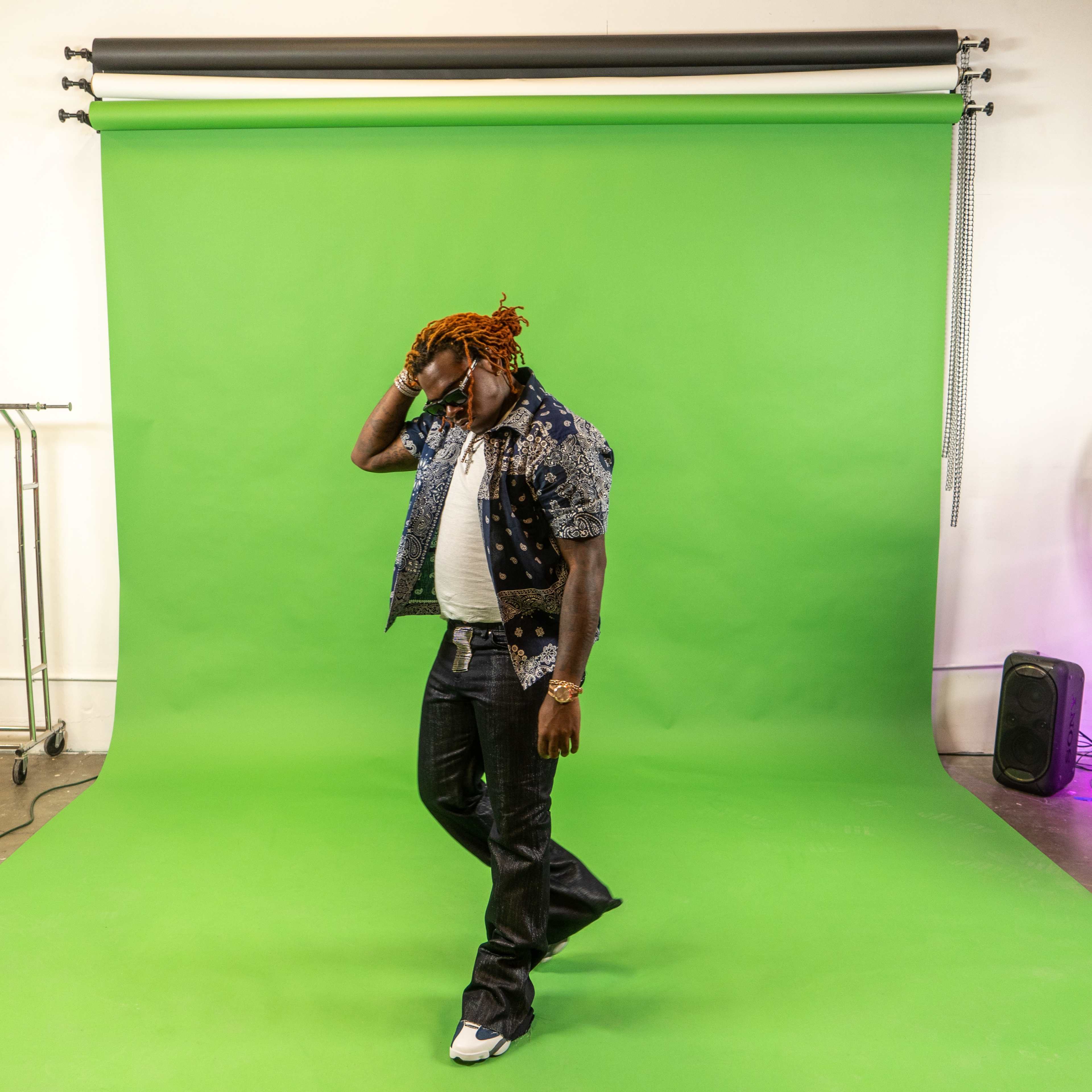 A person with orange hair walks in front of a green screen, wearing a patterned shirt and jeans.