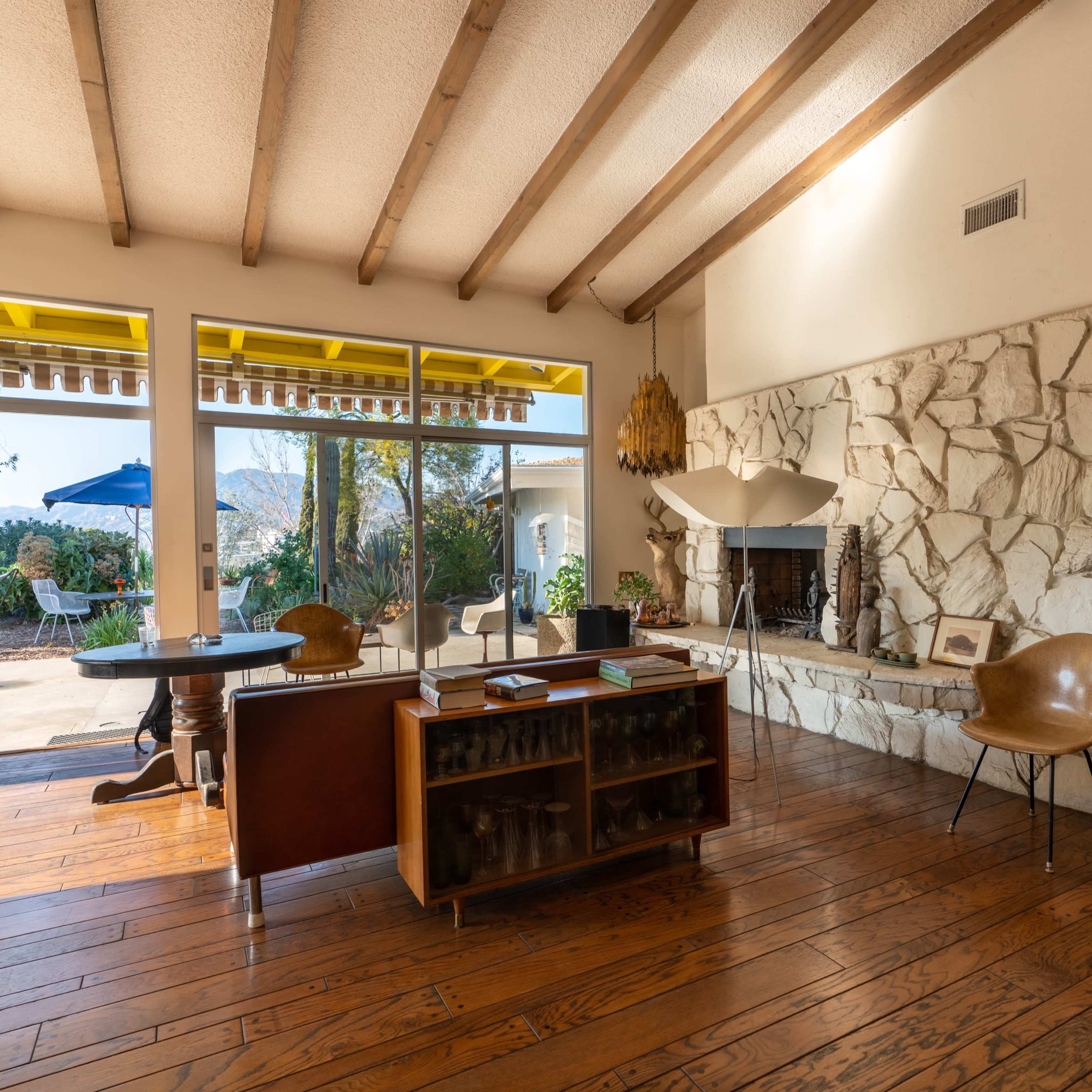 A modern living room with wooden beams, a stone fireplace, and a view of an outdoor patio through large windows.
