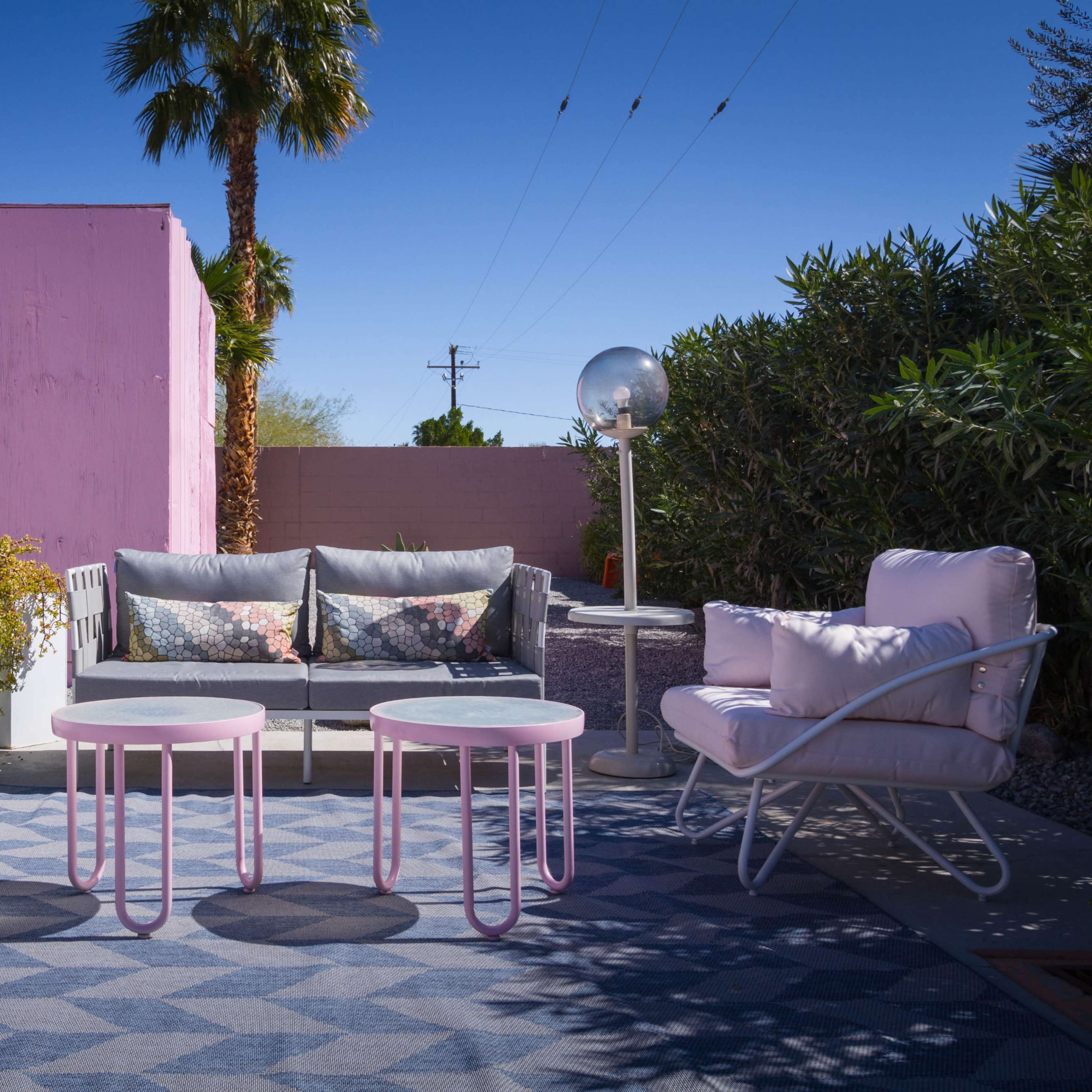 A cozy outdoor seating area featuring two pink armchairs, a gray sofa with decorative cushions, and two round tables, all set against a backdrop of palm trees and a pink wall.