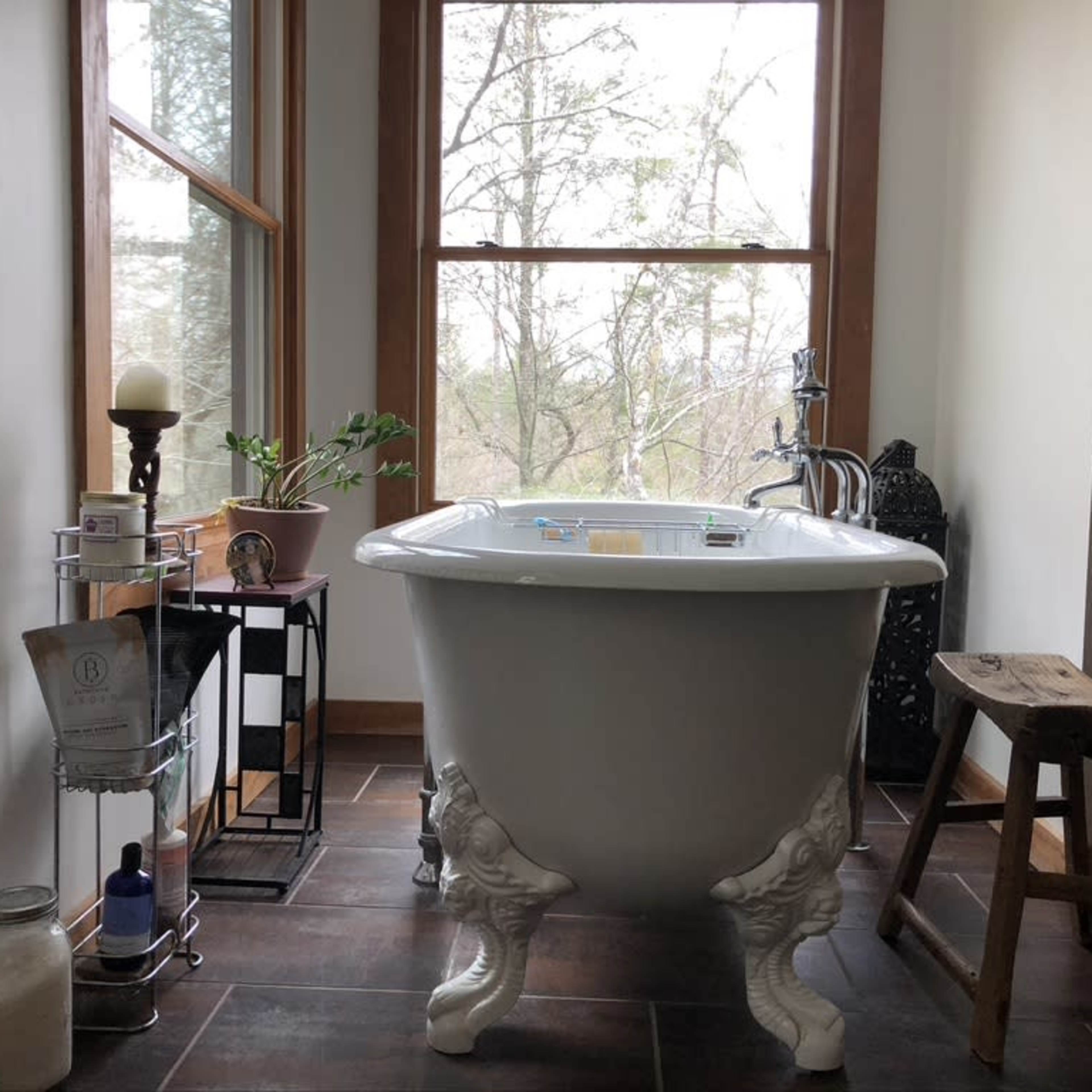 A freestanding clawfoot bathtub is positioned between large windows, with a small plant and various bathroom accessories nearby.