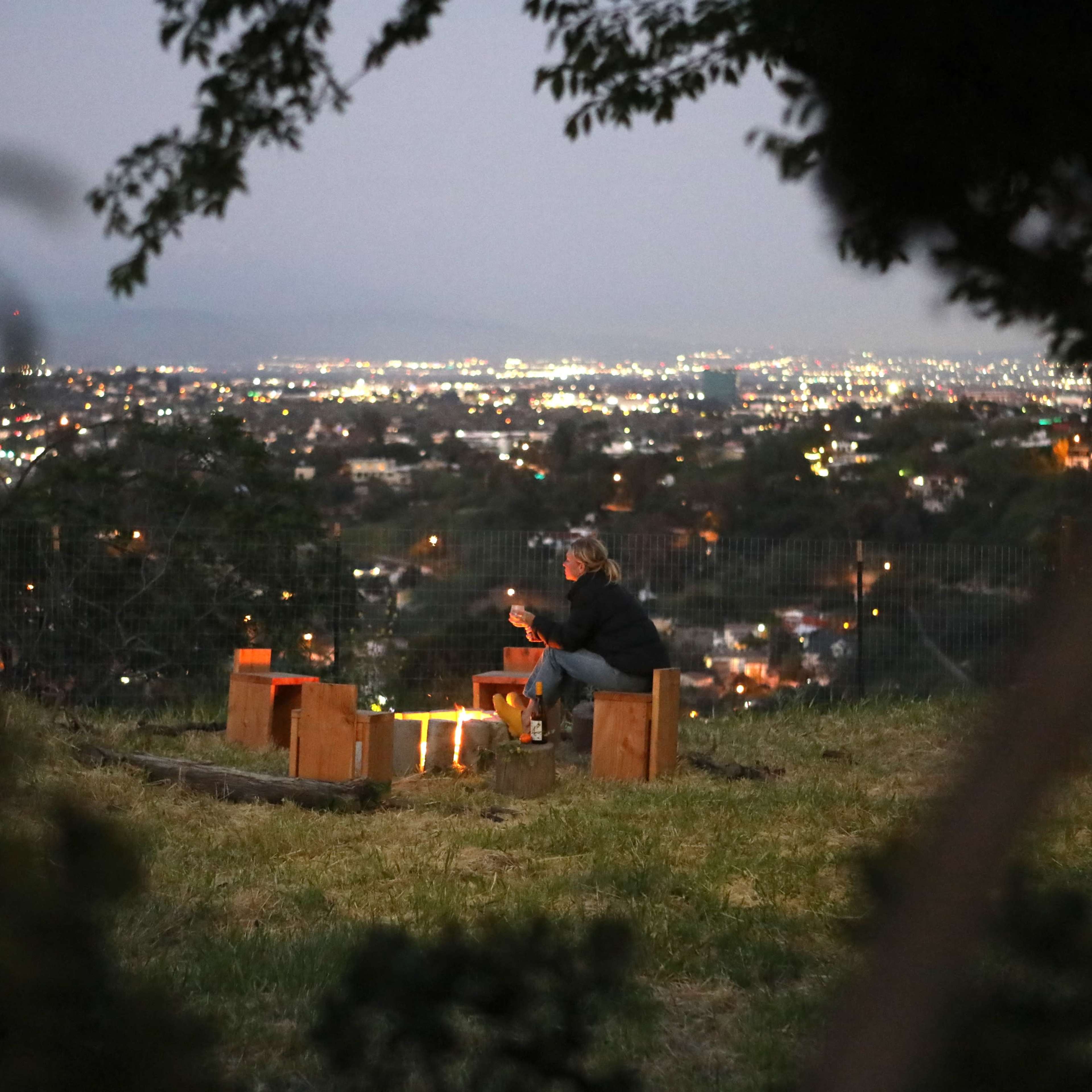 A person sits on benches around a campfire overlooking a city illuminated at night.