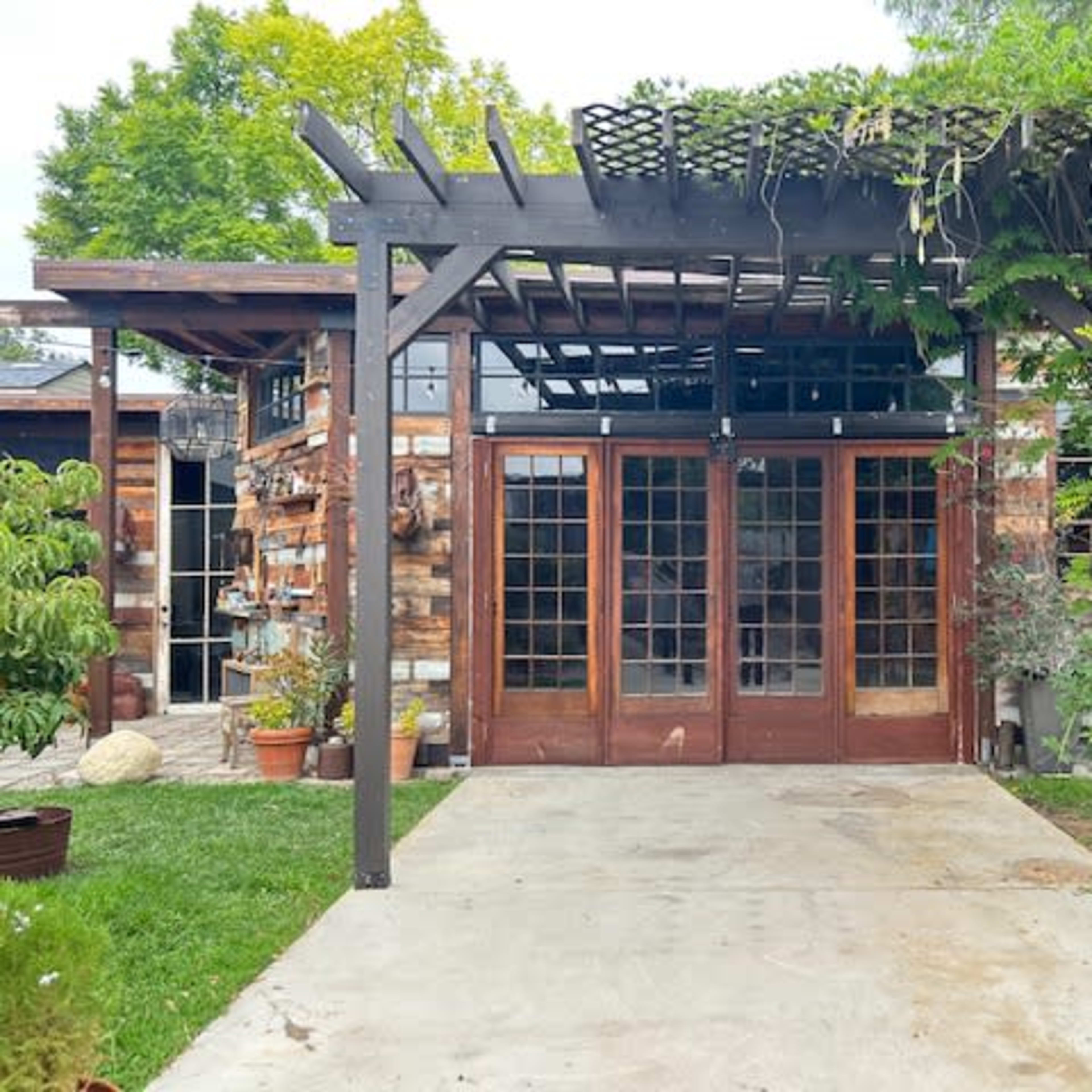 A rustic wooden structure with large glass doors, surrounded by greenery and a pergola with climbing plants.