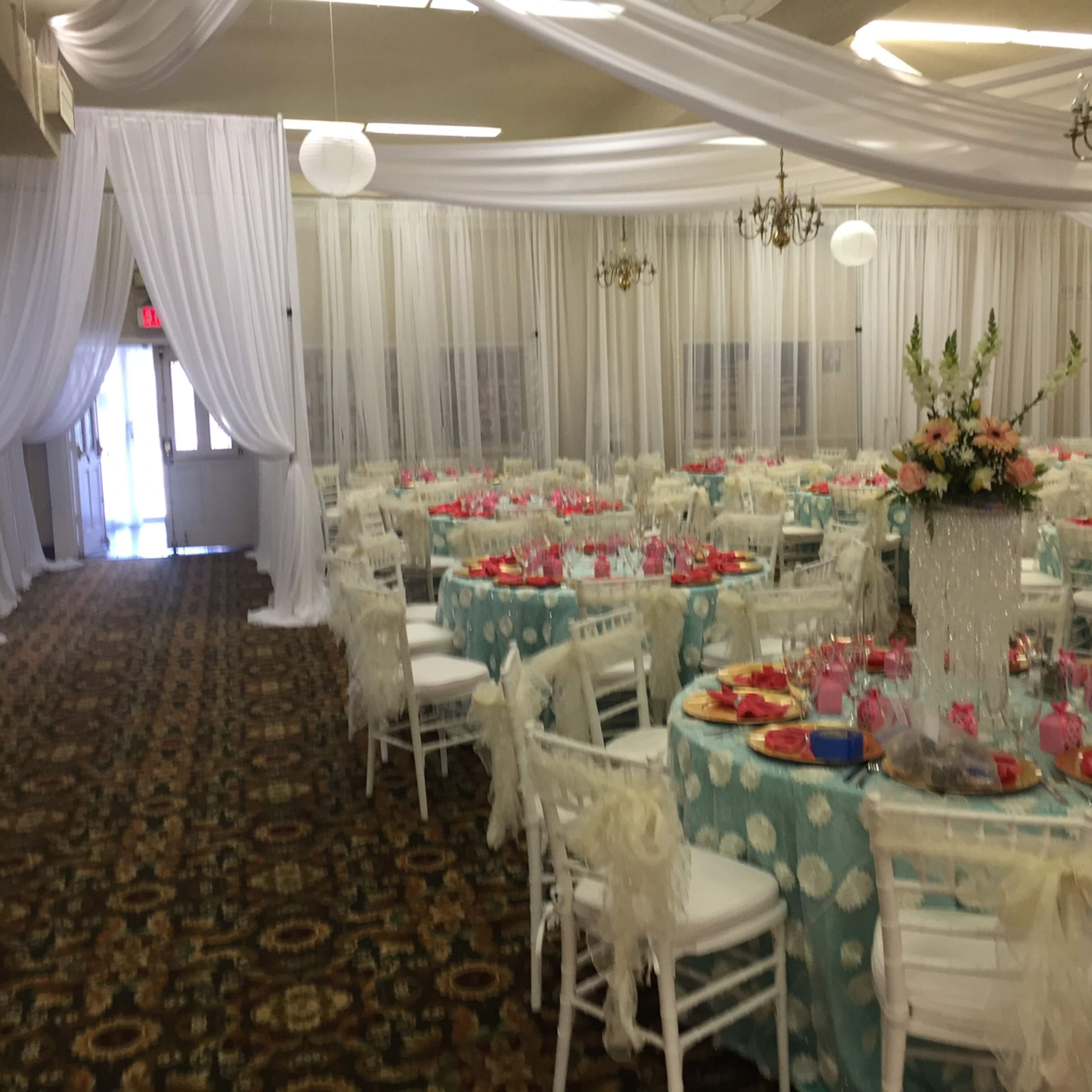 The room features elegantly set round tables with light blue tablecloths and white chairs, decorated with floral centerpieces and pink tableware, all under draped white fabric.