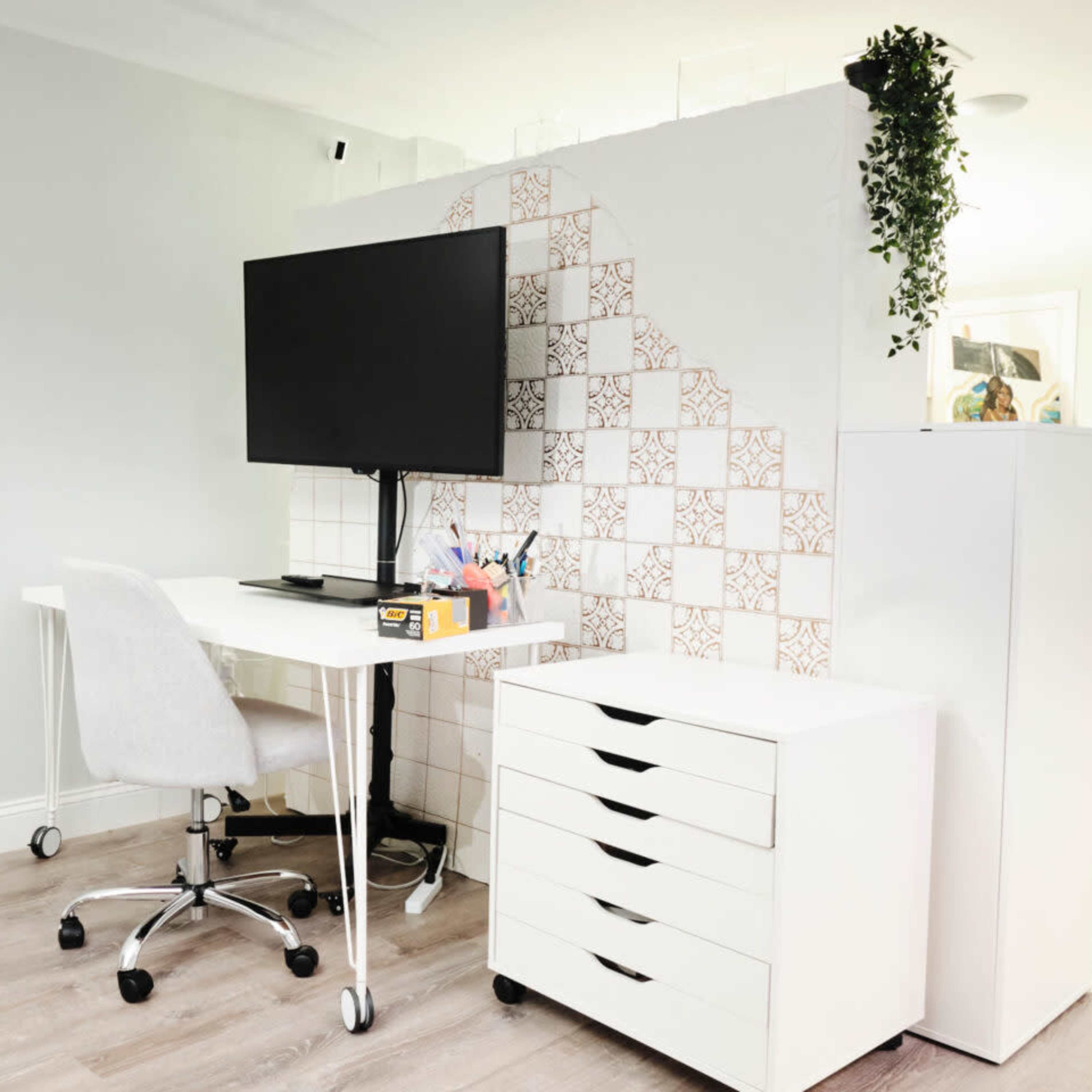 A modern office space featuring a desk with a monitor, a rolling chair, and a white storage unit against a partially tiled wall with a plant.