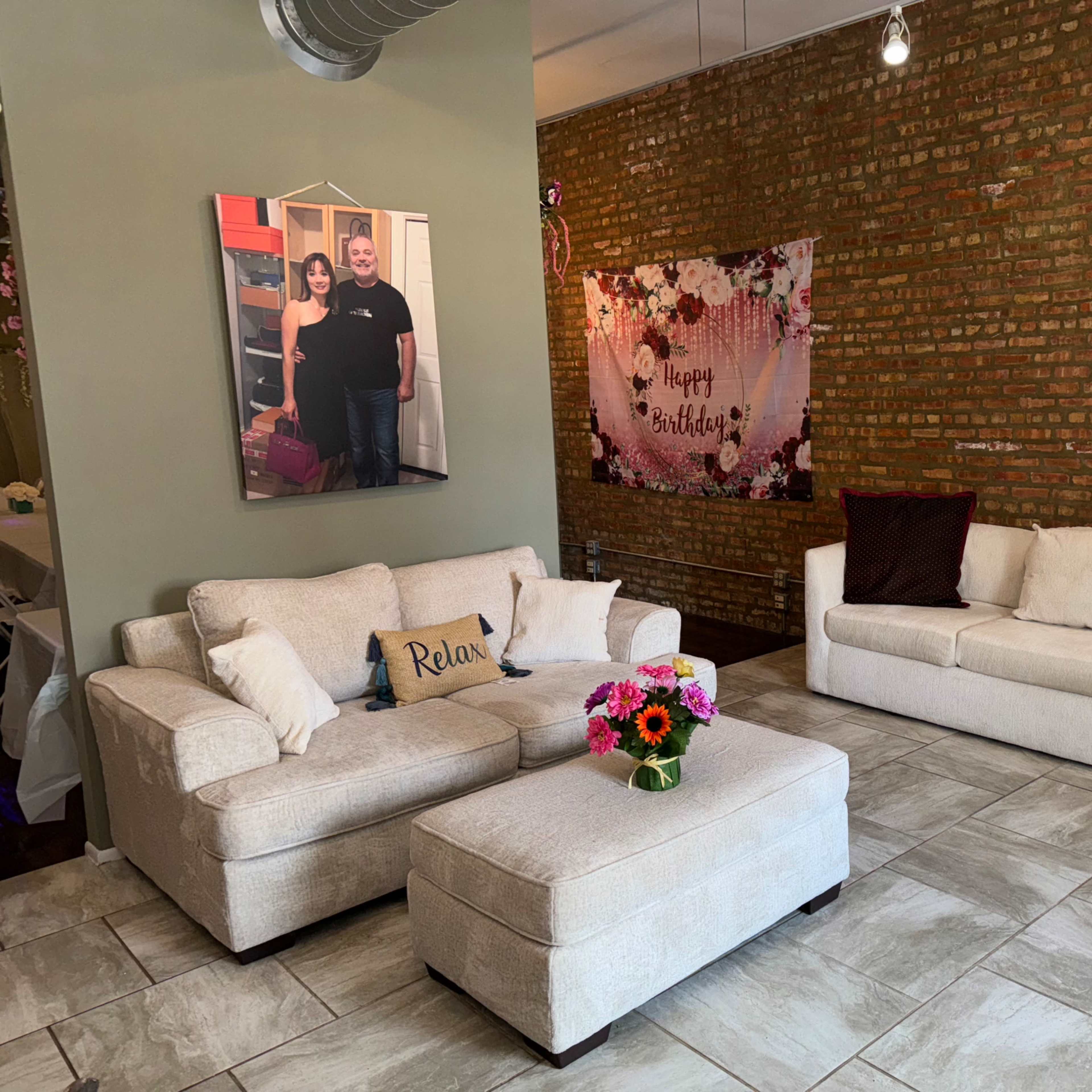 A modern living room with a light-colored sectional sofa and an ottoman, a colorful flower bouquet on the coffee table, and a birthday banner on the brick wall.