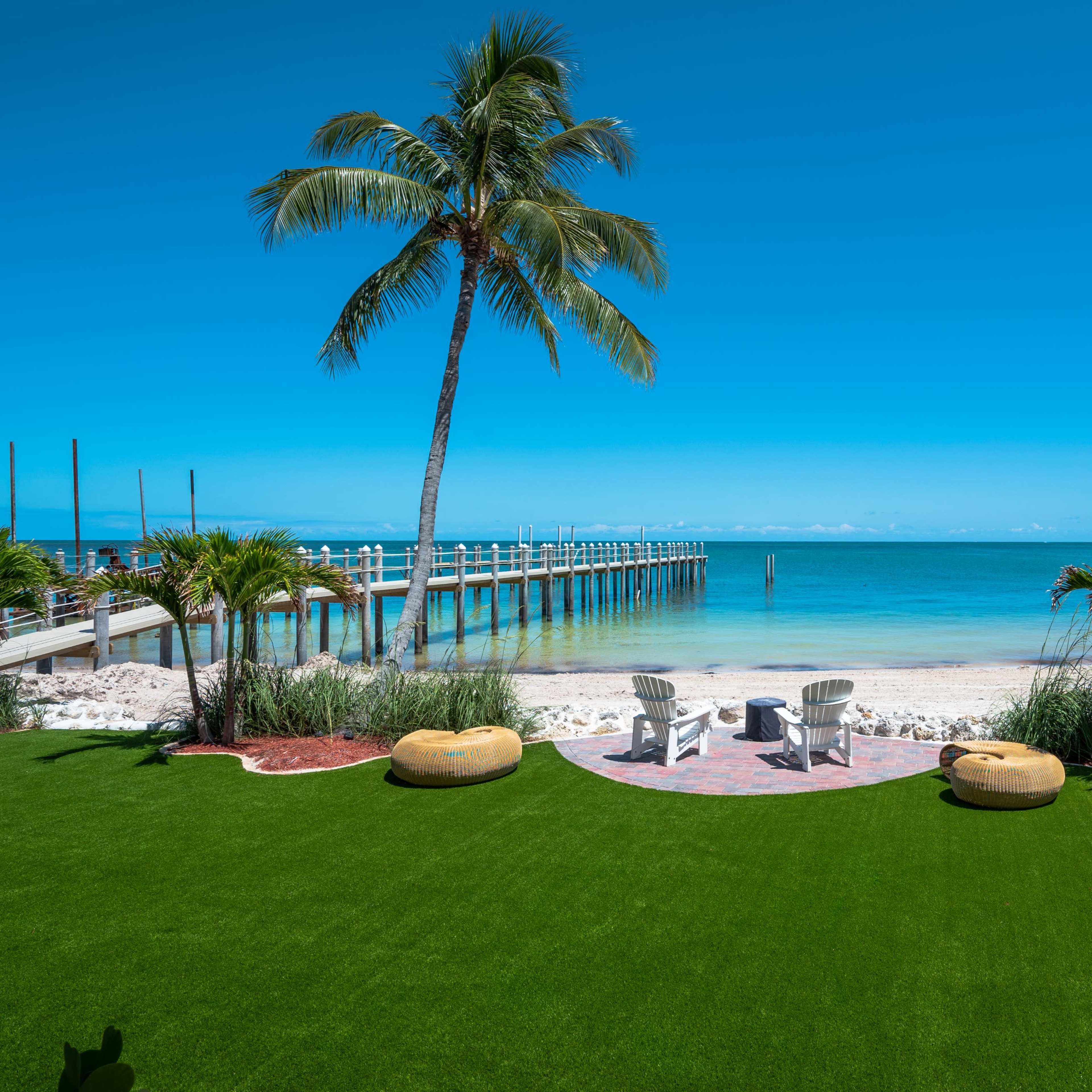 A sandy beach with a wooden pier and two chairs facing the ocean, framed by palm trees and green landscaping.