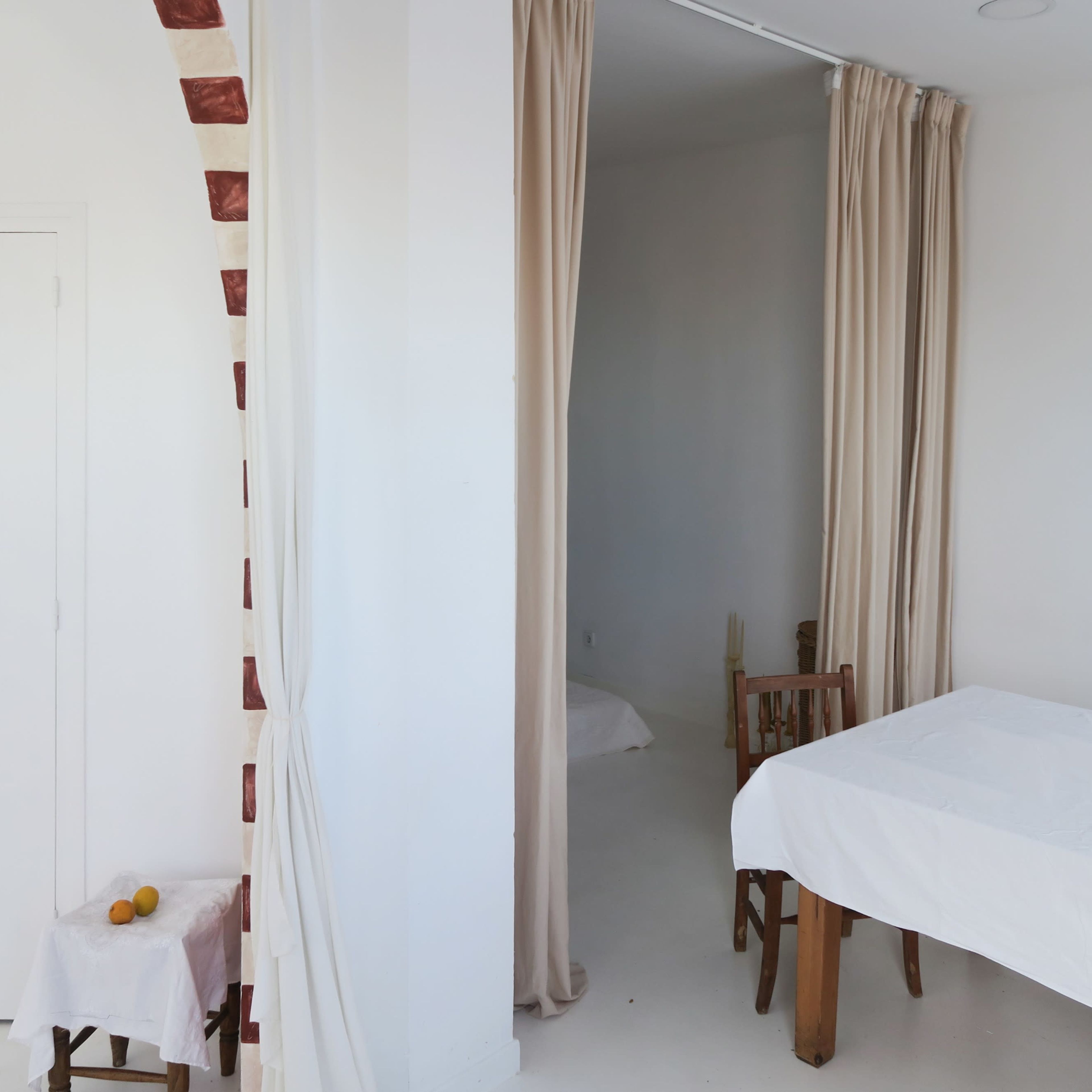 A bright, minimalistic interior with a white table and chairs, a doorway draped with curtains, and a wall partially covered with a brick arch.