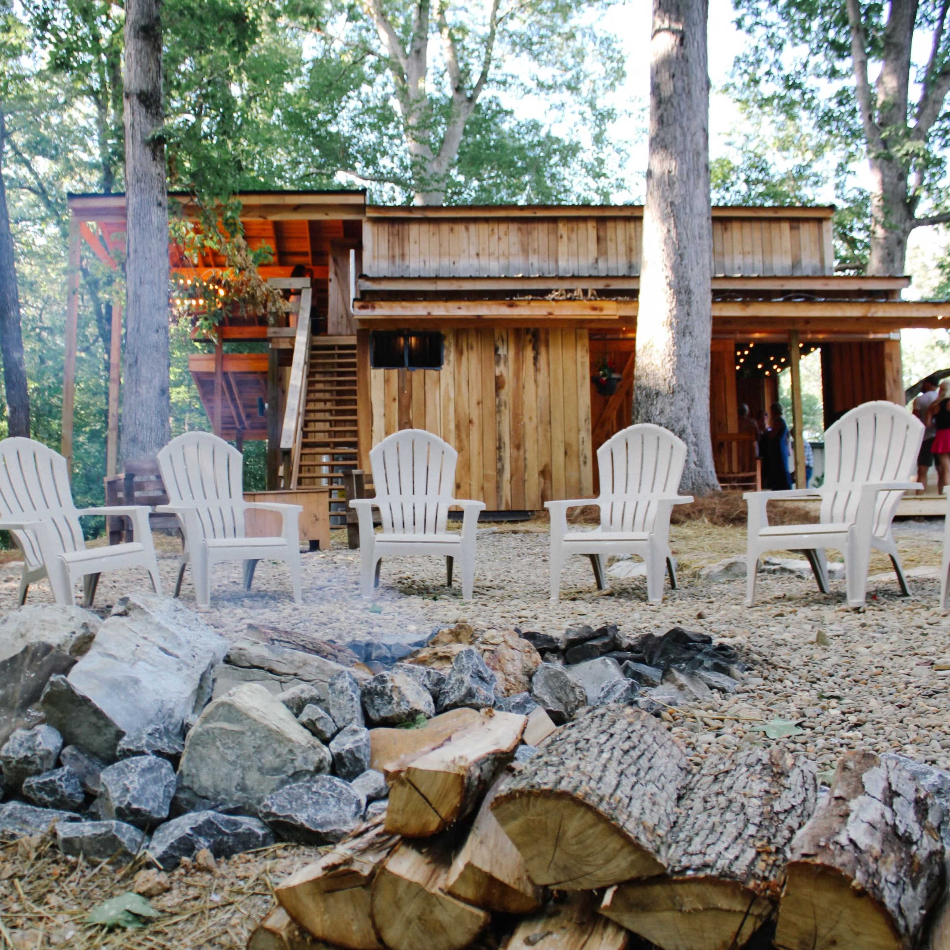 A rustic wooden cabin surrounded by trees features a fire pit with several white chairs arranged in a circular formation.