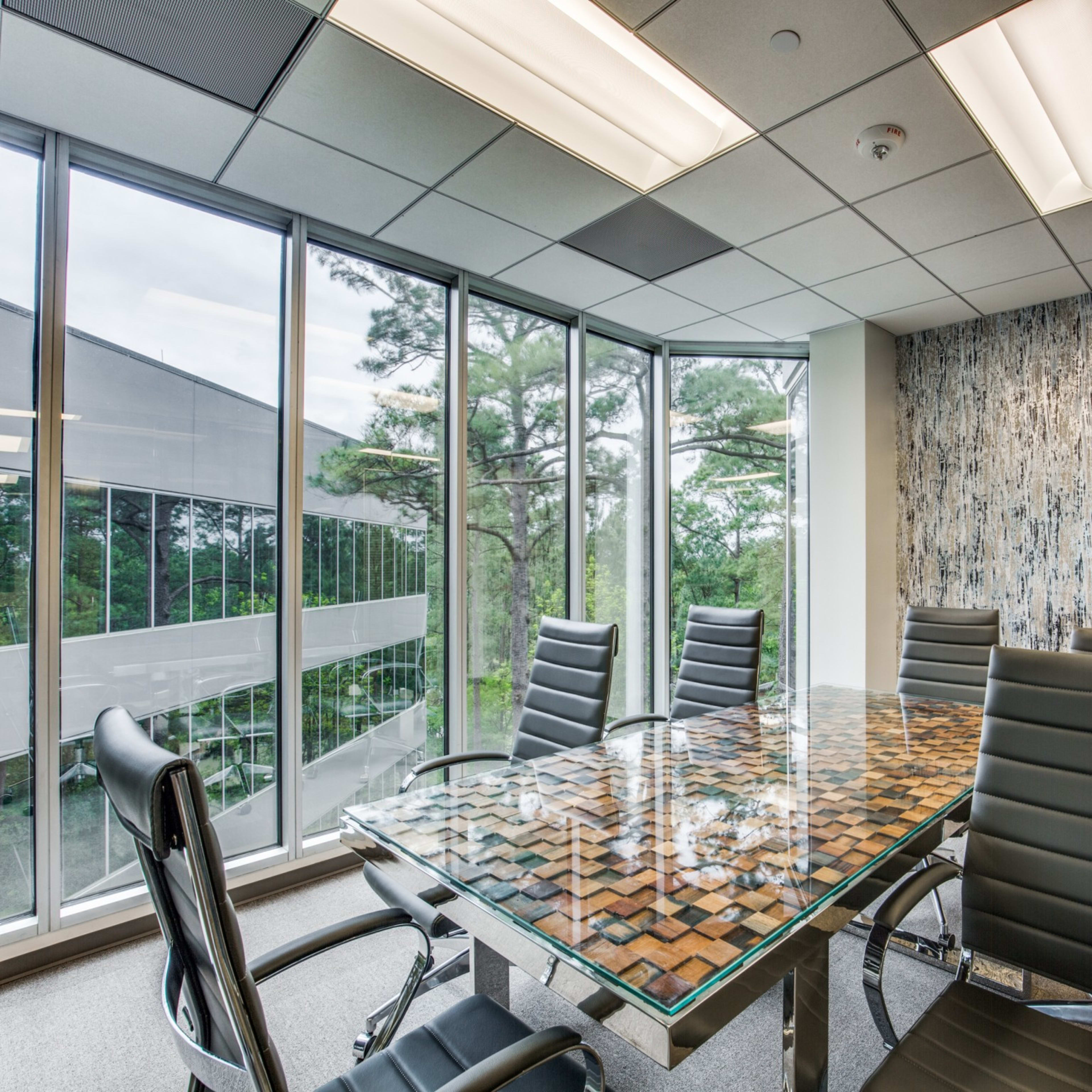 A conference room features a glass-topped table surrounded by black leather chairs, with large windows displaying a view of trees.