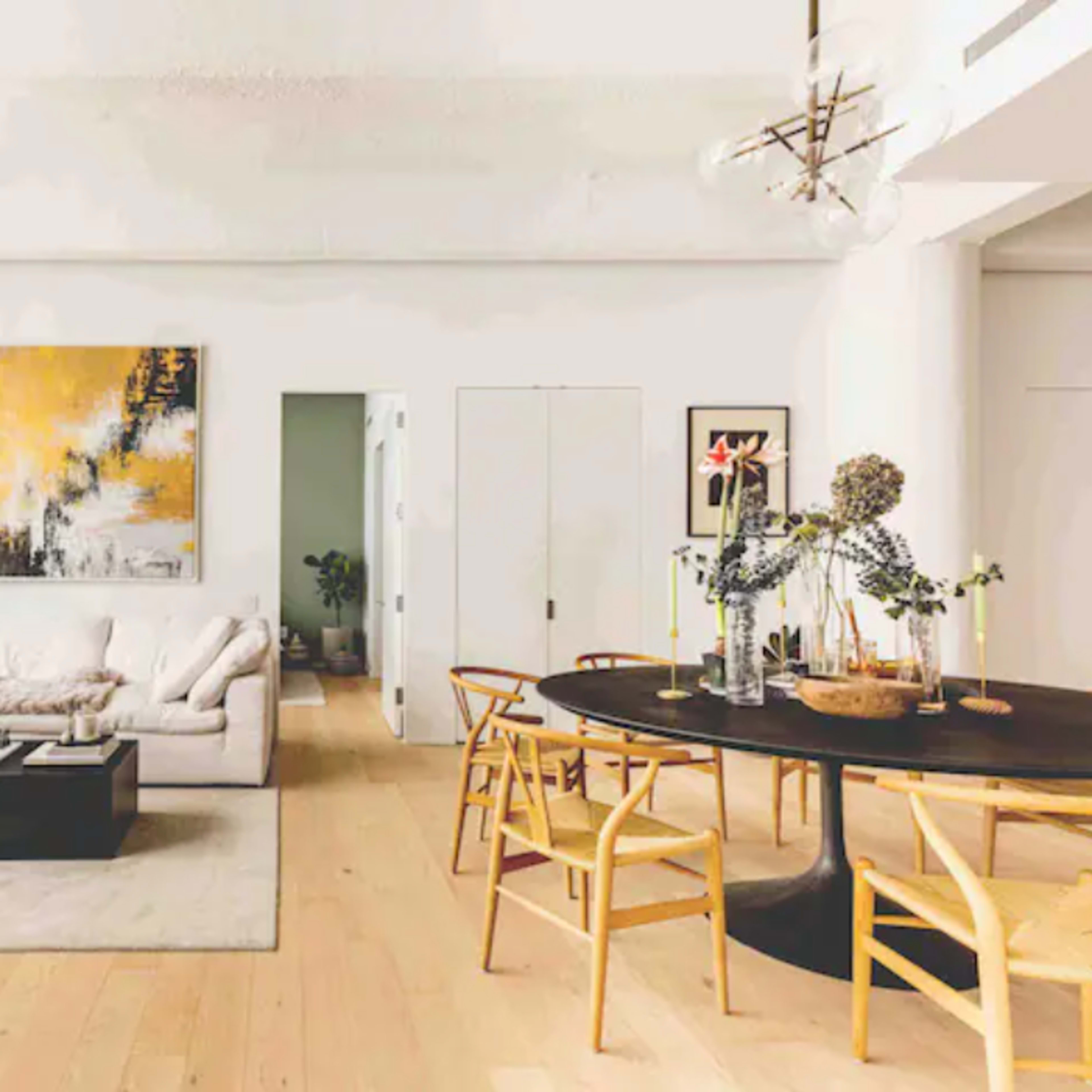 A modern living room features a light-colored sofa, a black coffee table, and a dining area with a round table surrounded by chairs, illuminated by large windows.