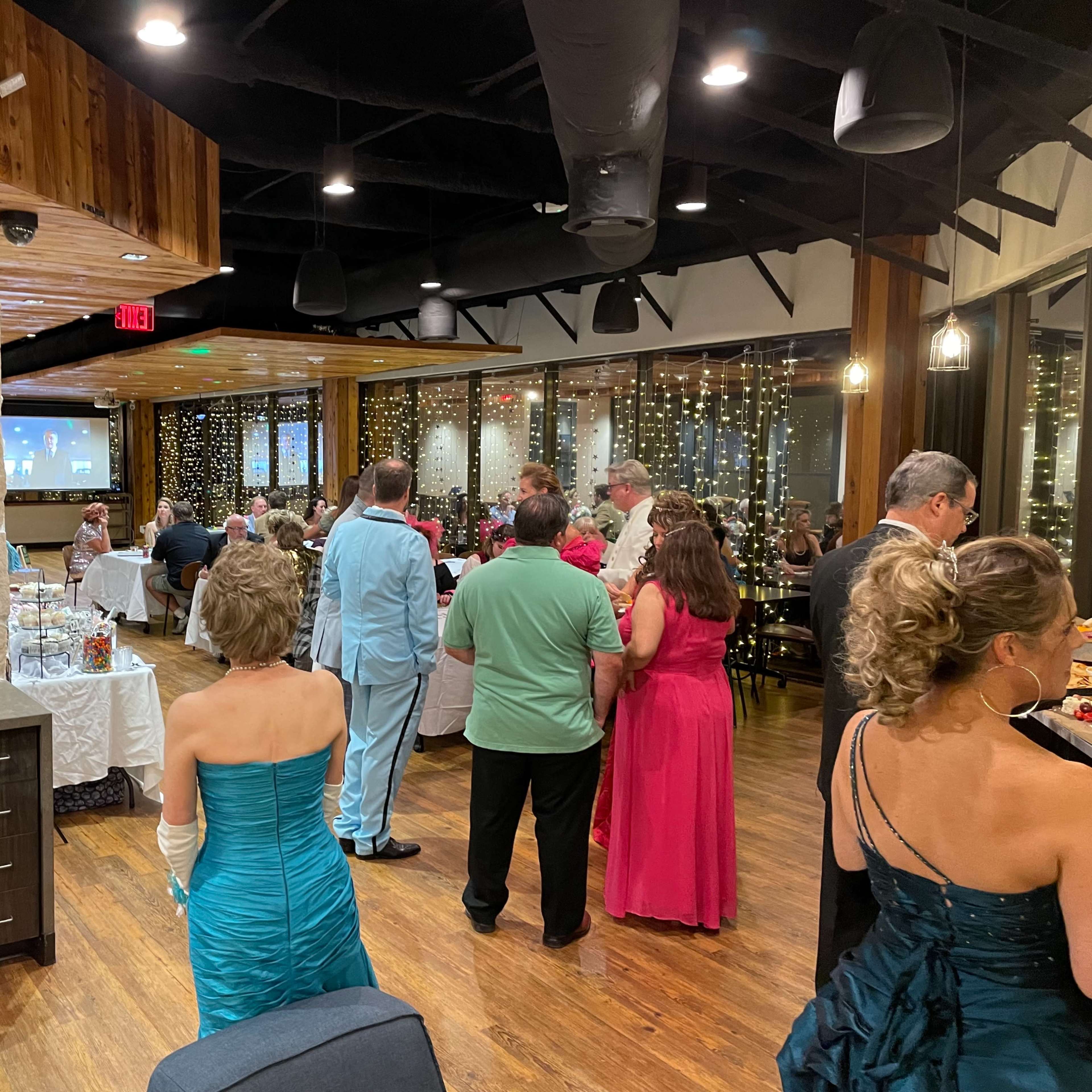 A group of attendees in formal attire socialize at a well-lit indoor event with a stone fireplace and string lights in the background.