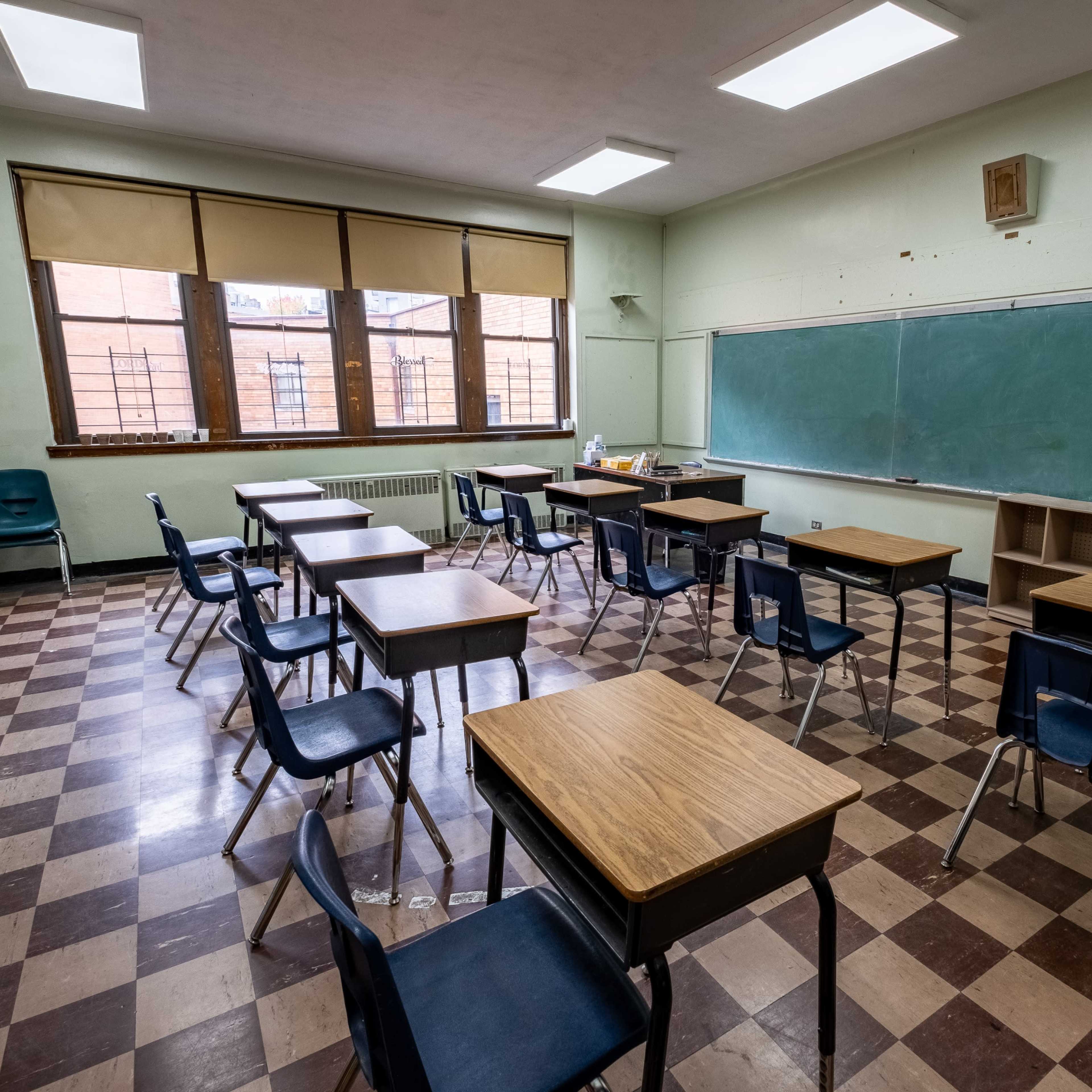 A classroom with wooden desks and blue chairs arranged around a checkered floor, featuring a chalkboard and windows lined with blinds.