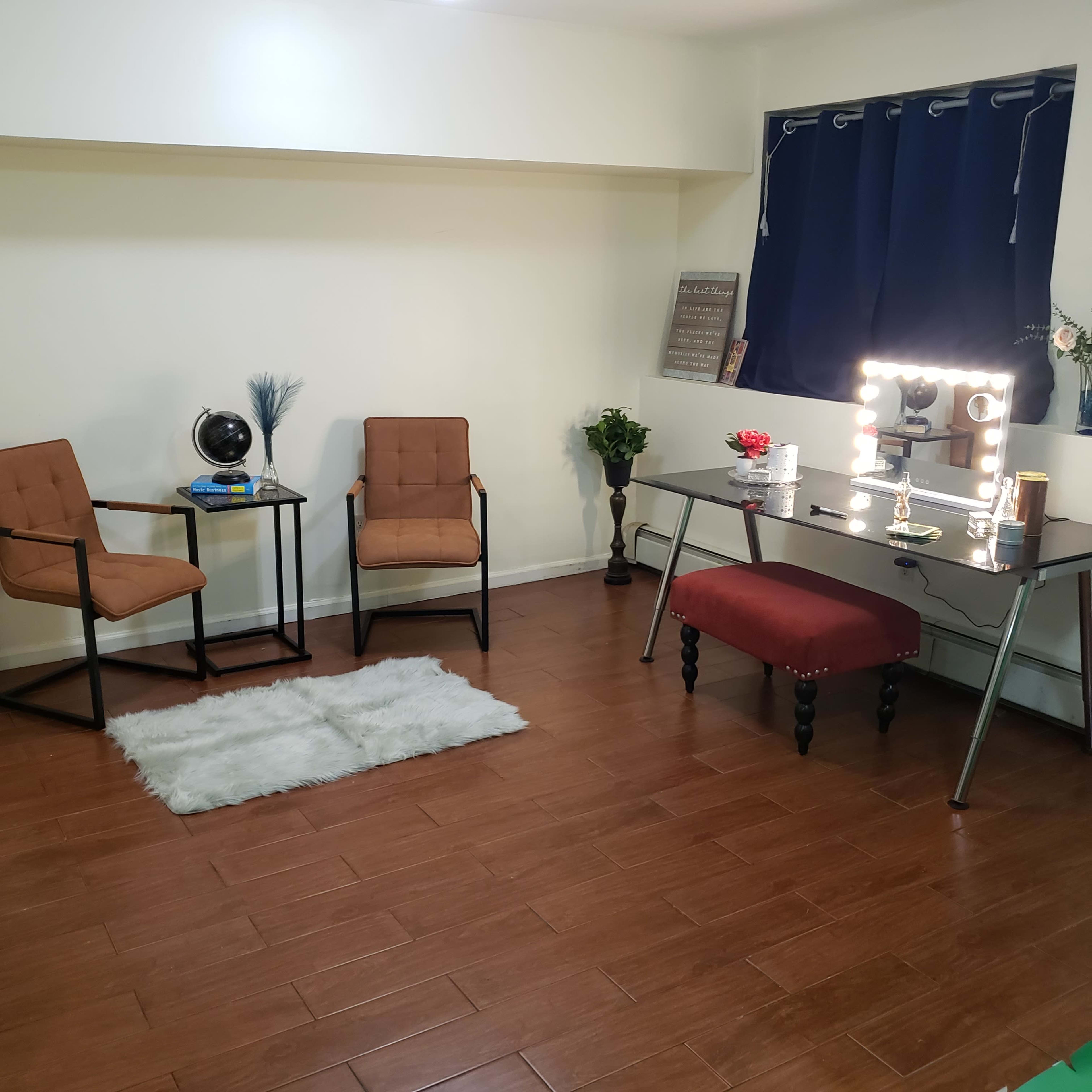A minimalist room with two brown chairs, a vanity table featuring a large mirror and light bulbs, and a decorative accent rug on a wooden floor.