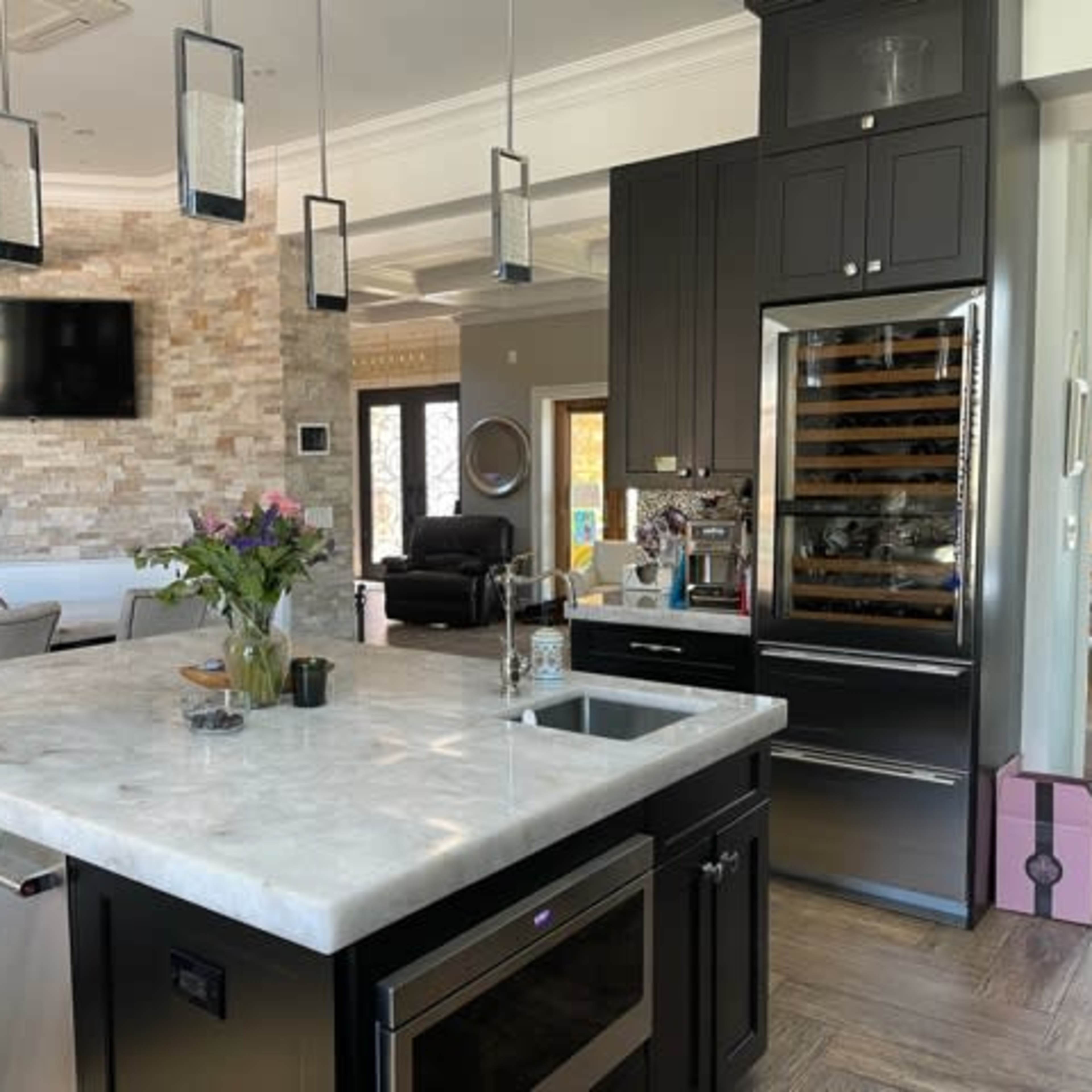 A modern kitchen featuring a large marble island with a sink, black cabinetry, and a wall-mounted flat-screen TV.