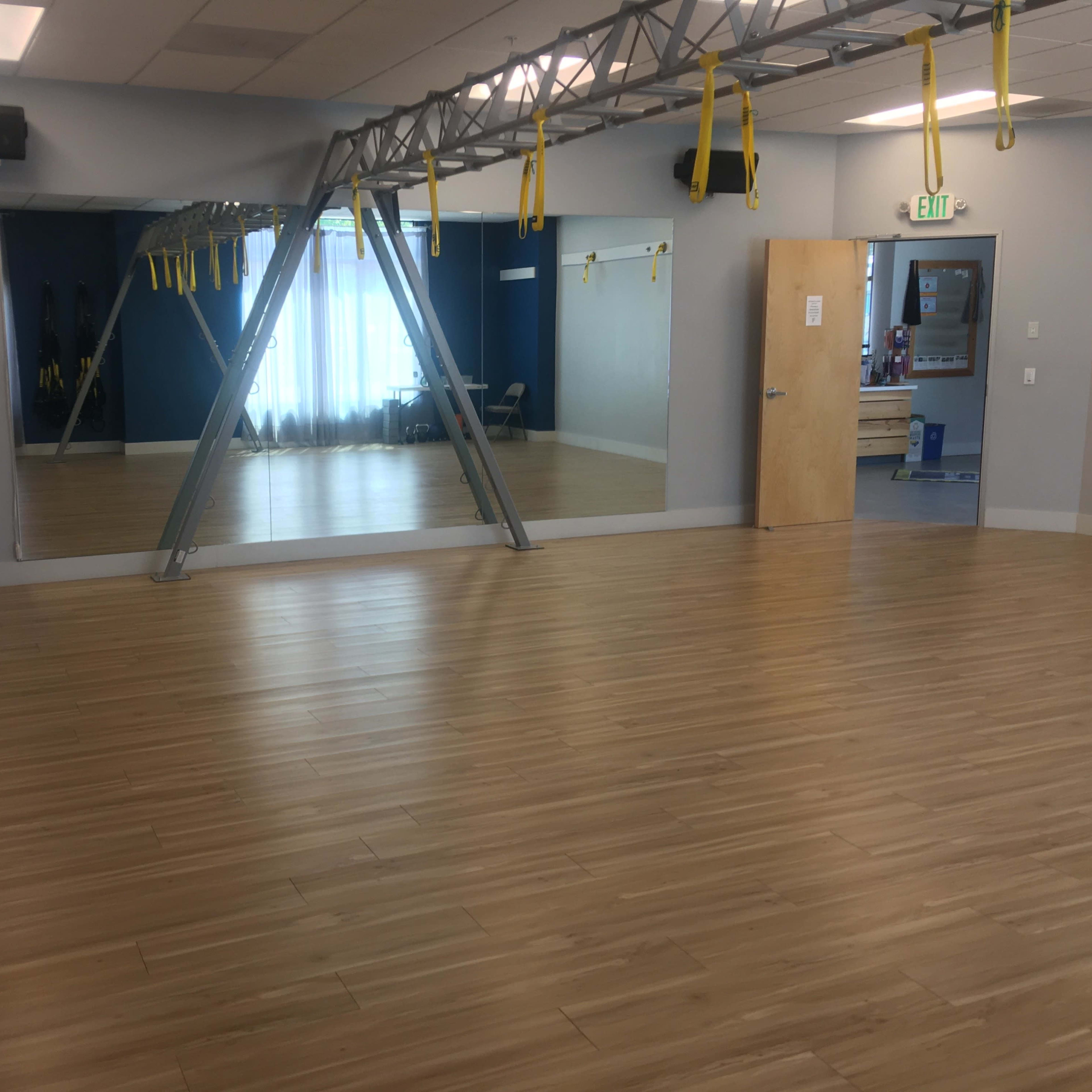 A spacious exercise studio with wooden flooring, equipped with suspension training straps, large mirrors, and natural light filtering through windows.