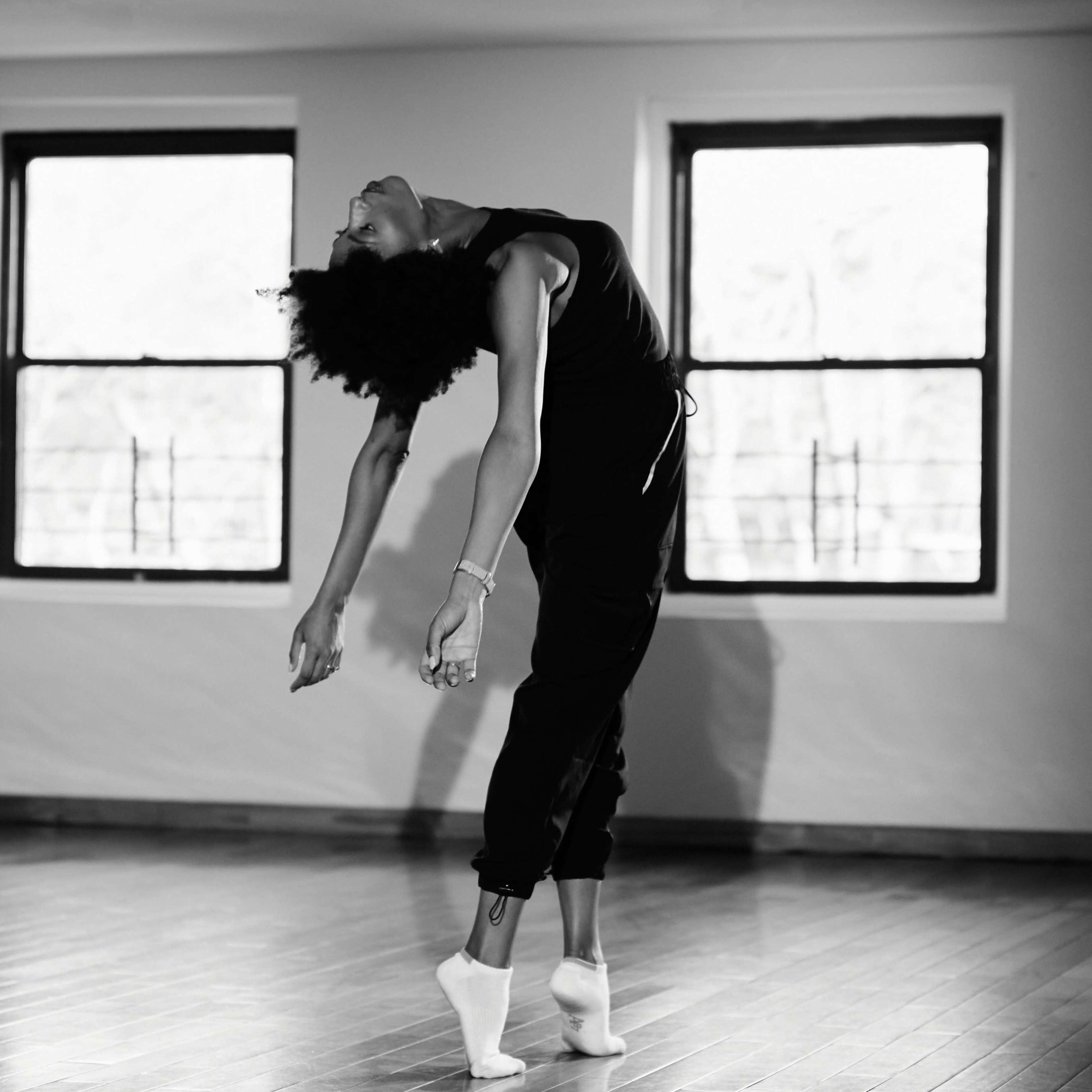 A dancer in a black outfit leans backward with arms extended in a sunlit studio with wooden floors and large windows.