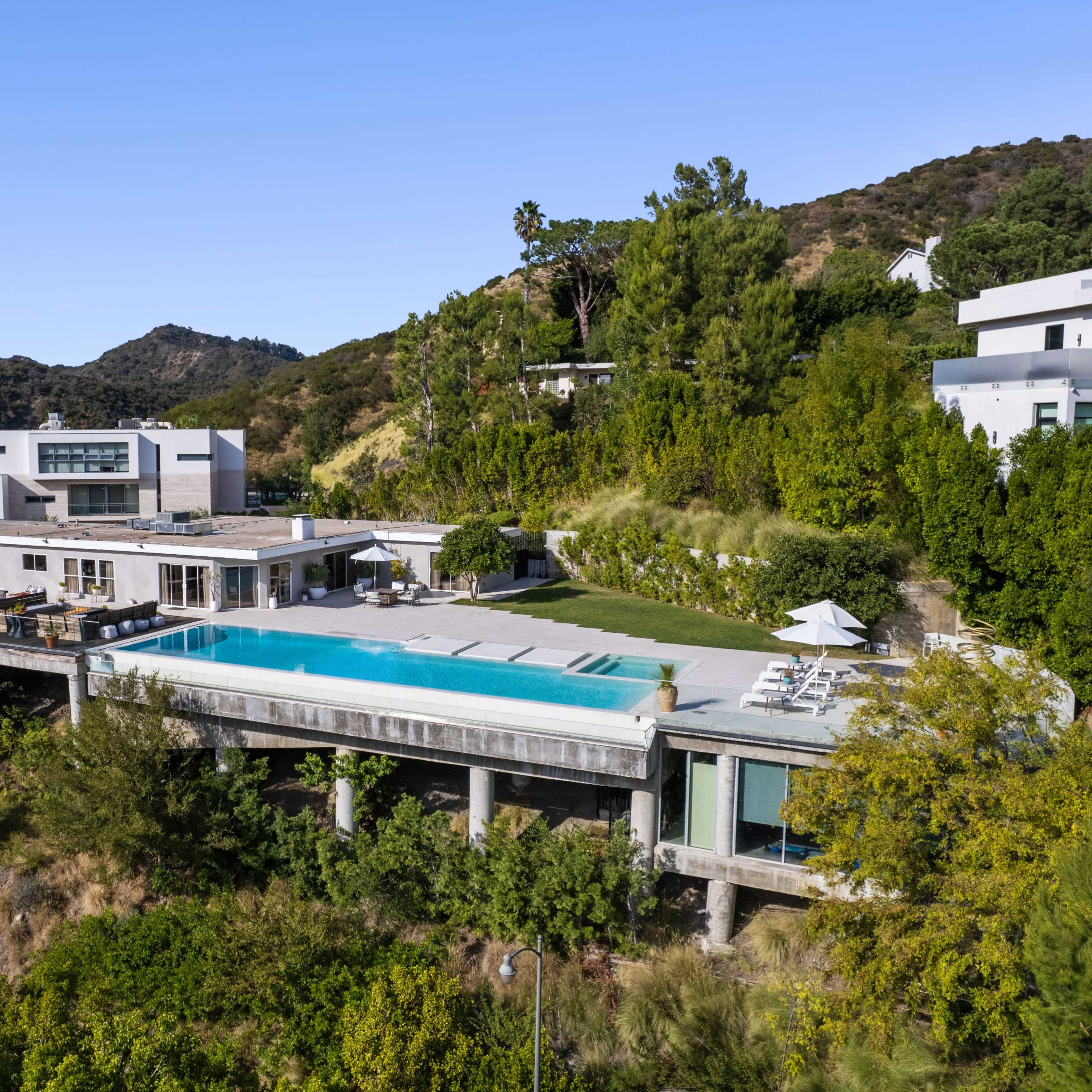 A modern house with a large swimming pool set on a hillside surrounded by greenery and mountains.