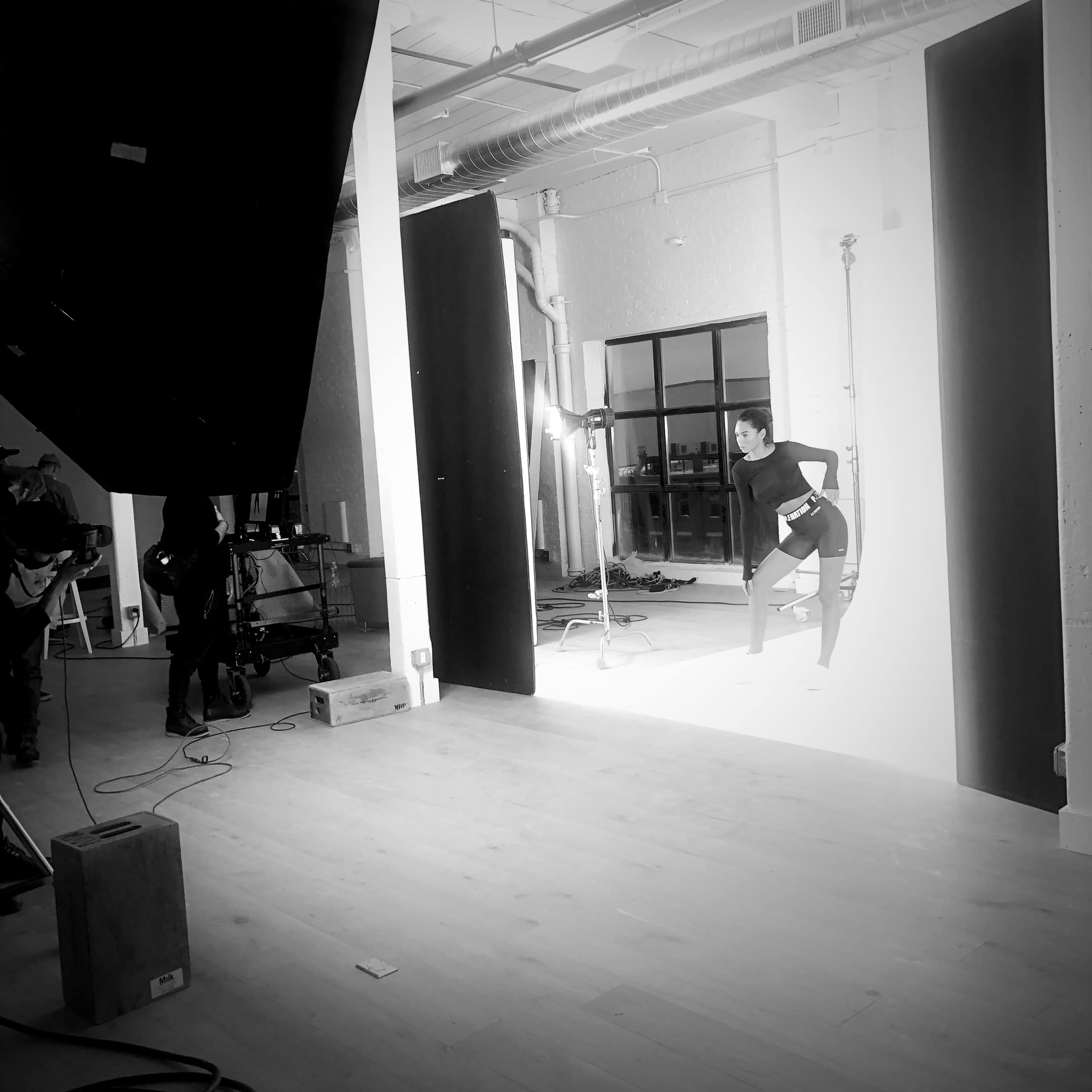 A studio space features a model posing against a backdrop with large softboxes and equipment set up around her.