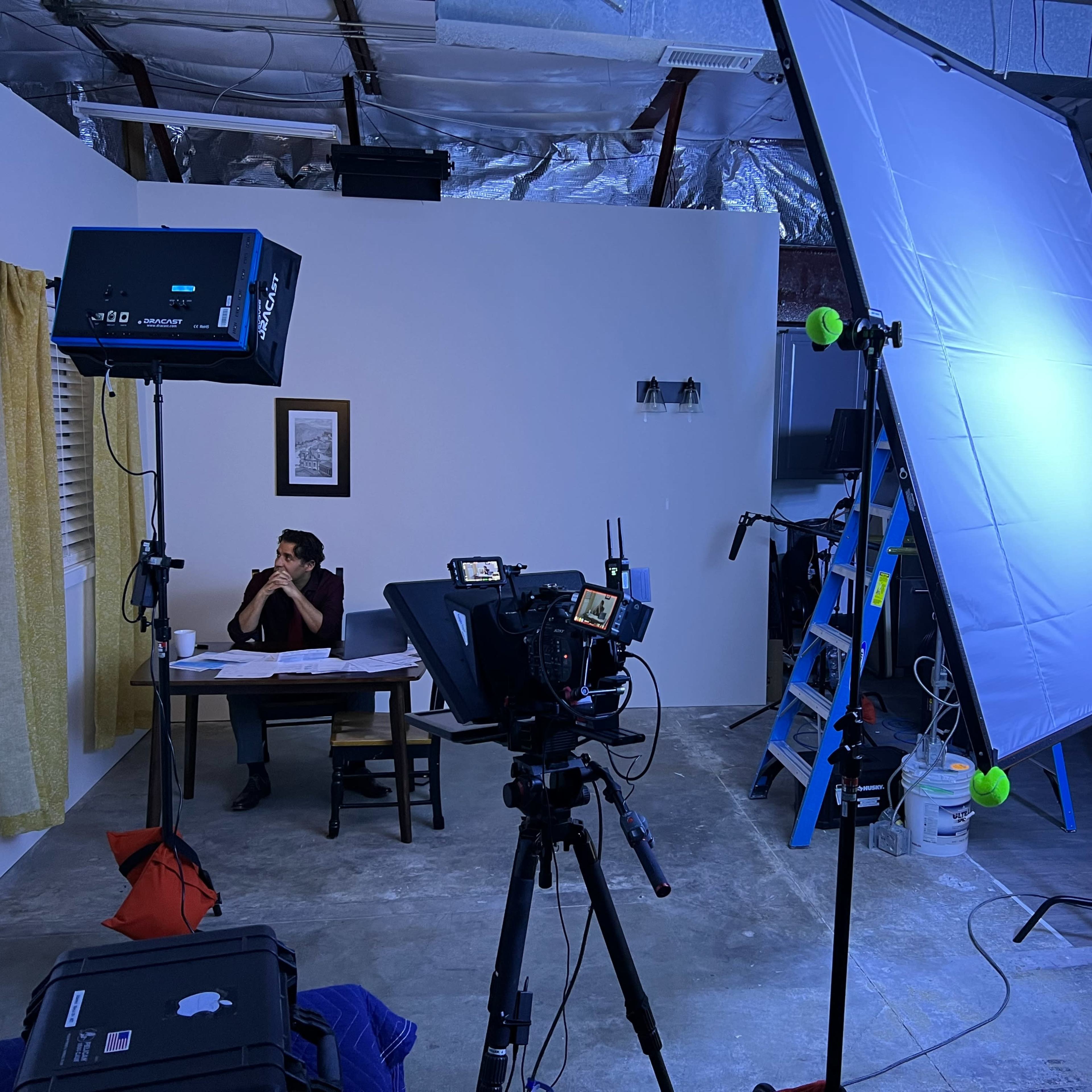 A film or video production setup with a person sitting at a table, cameras and lighting equipment arranged around them in a dimly lit room.