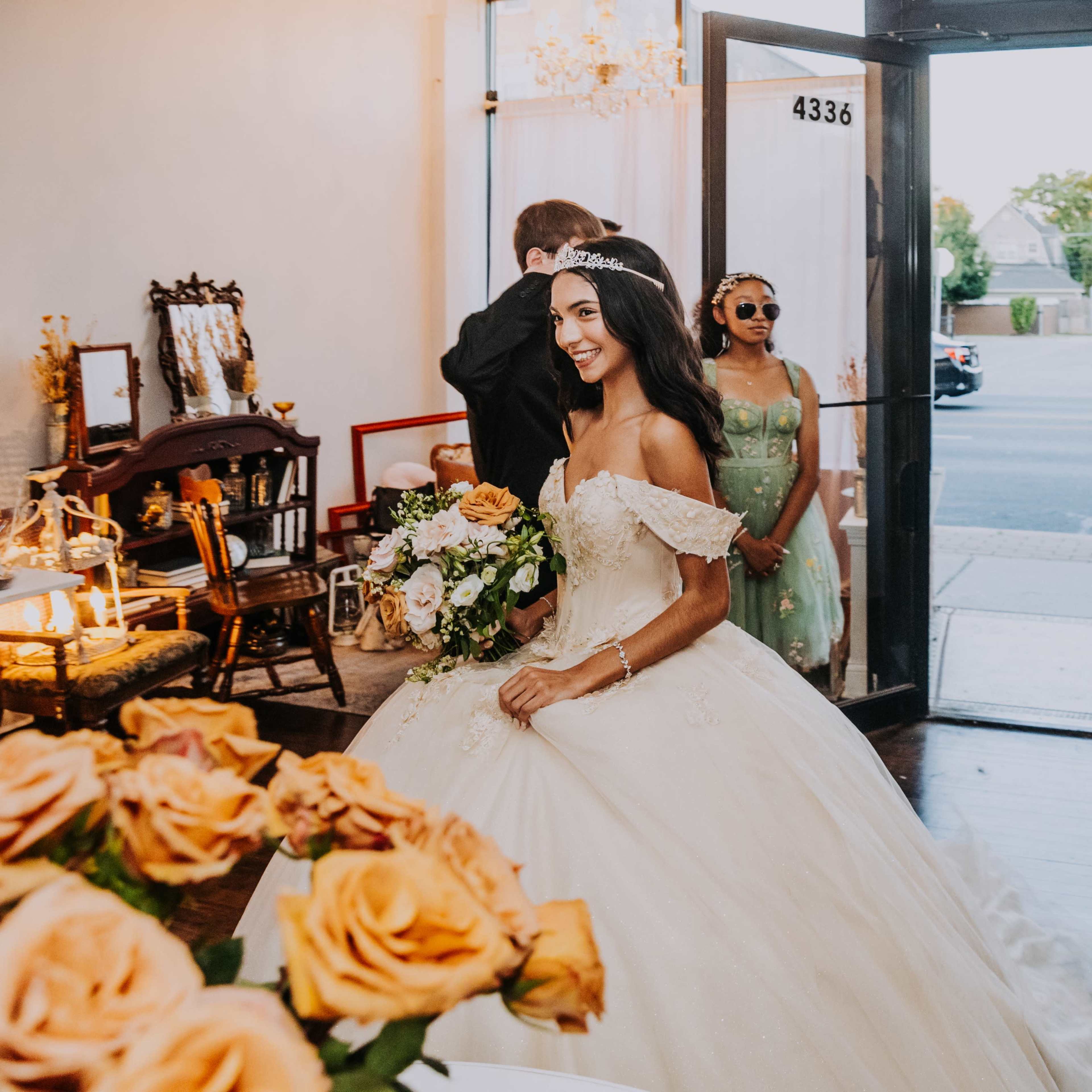 A bride in a white gown holds a bouquet of flowers while walking towards a group of guests in a decorated event space.