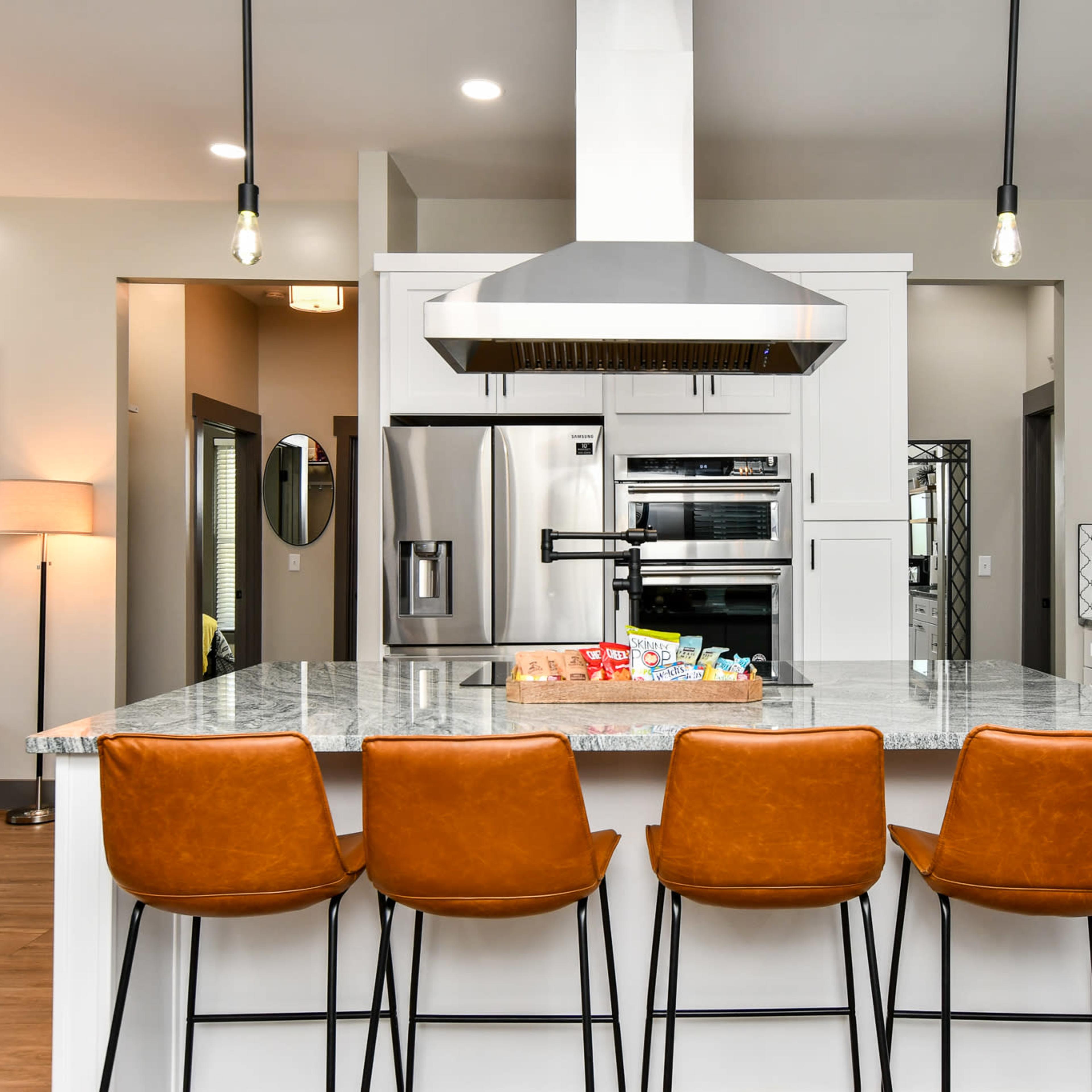 A modern kitchen features a large island with four brown bar stools, stainless steel appliances, and a hood above the stove.