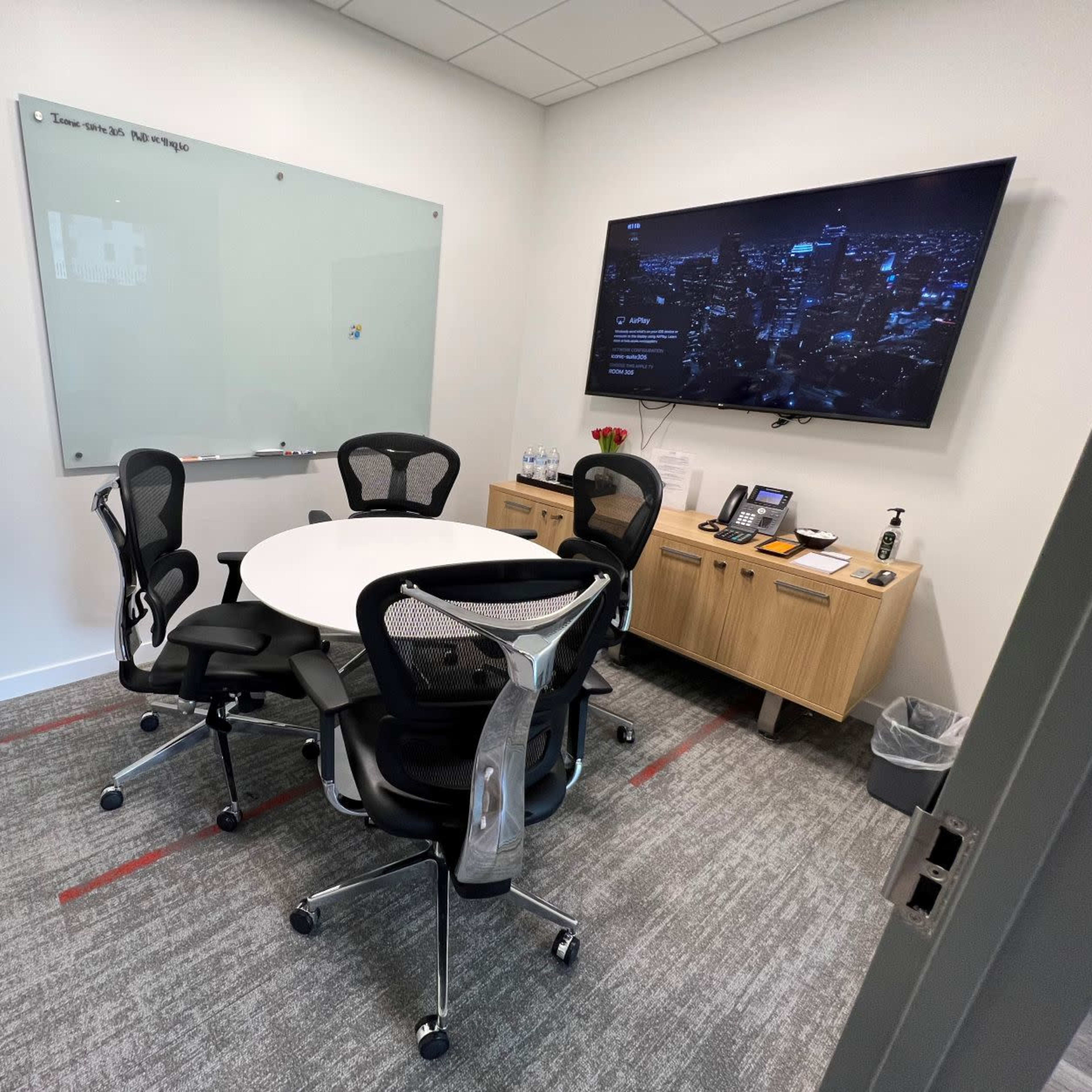 A small meeting room features a round table surrounded by six office chairs, a wall-mounted TV, a whiteboard, and a potted plant.
