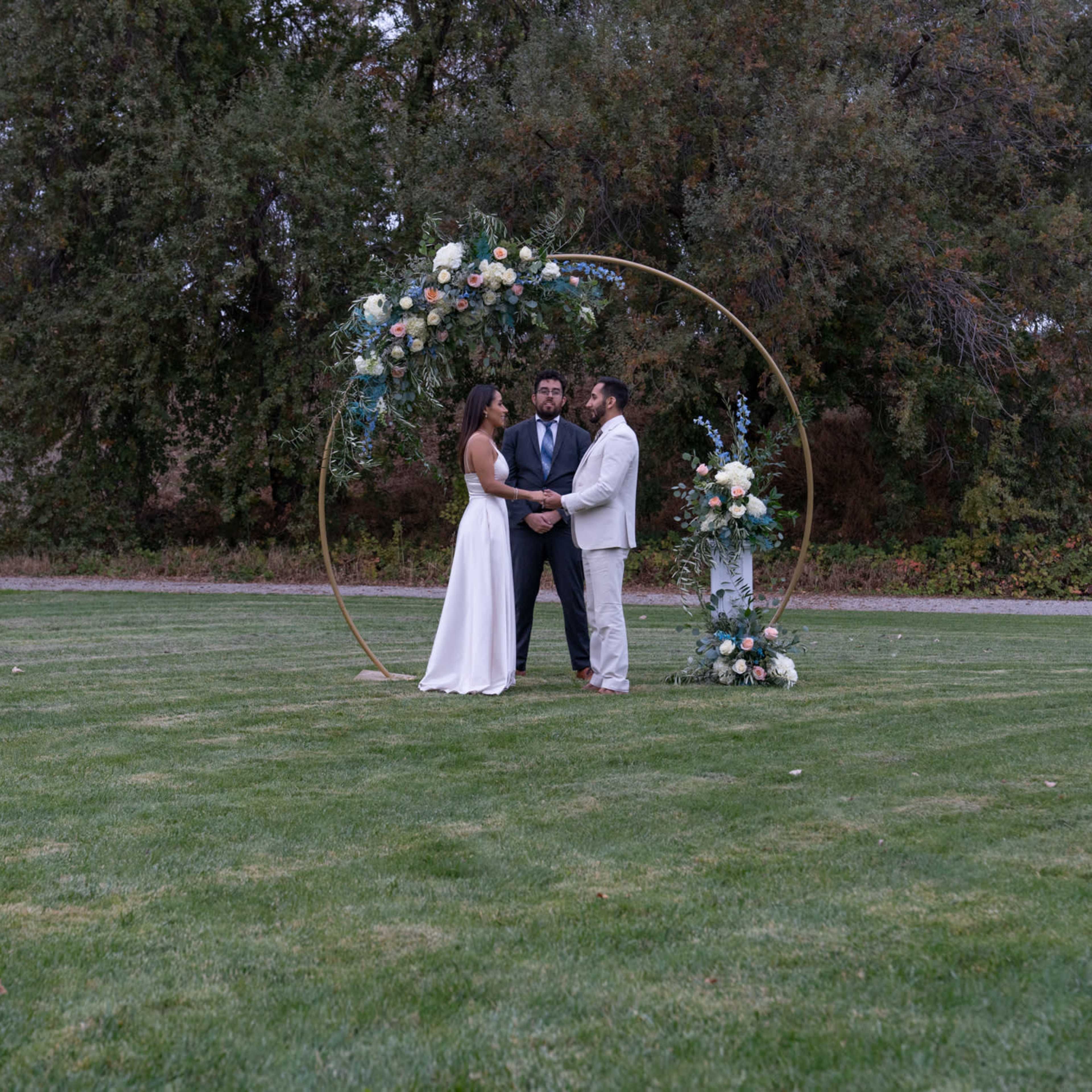 A couple stands under a floral arch while officiating their wedding ceremony on a grassy field.