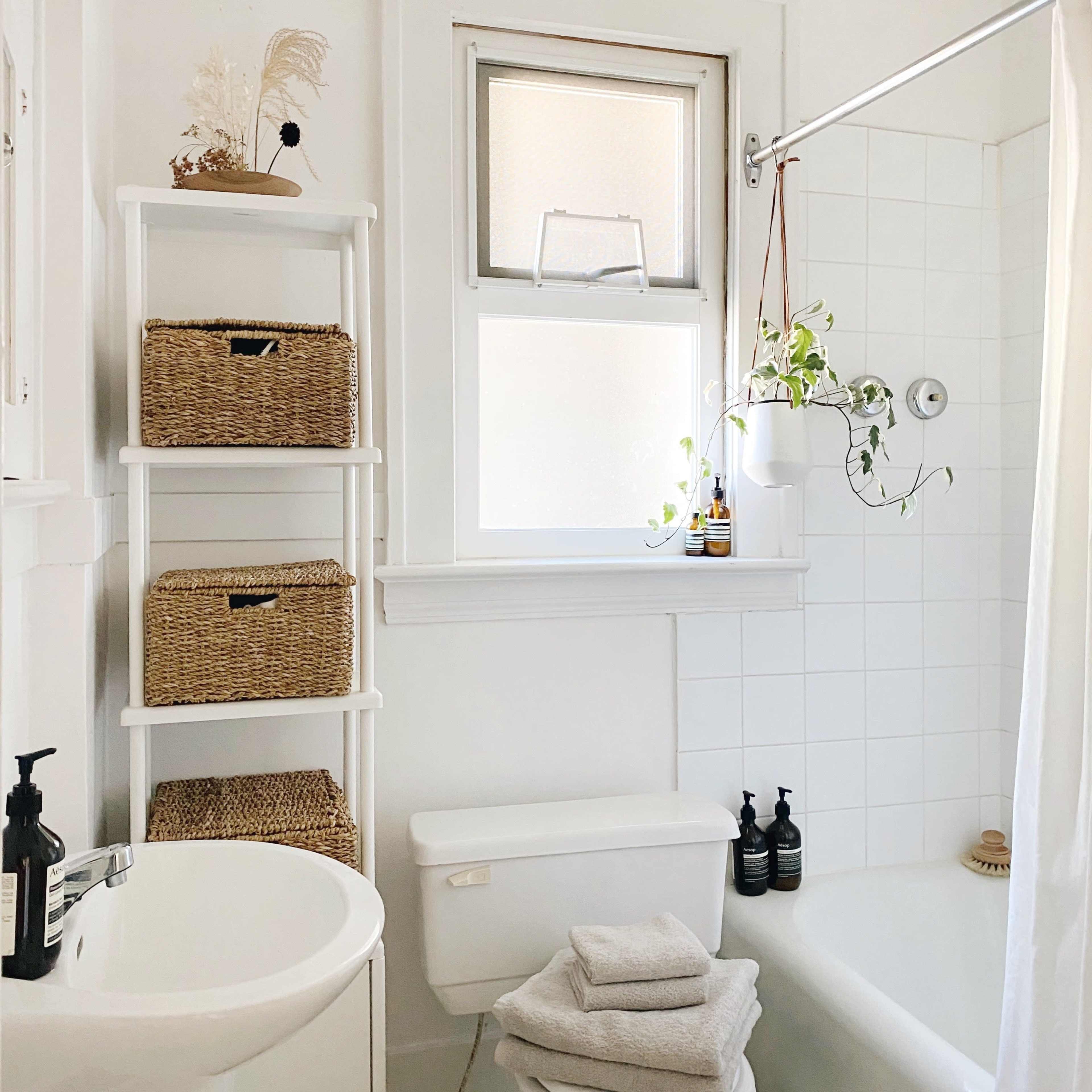 A bright bathroom features a tub, a white shower curtain, a small sink, and a shelf with wicker baskets and potted plants.