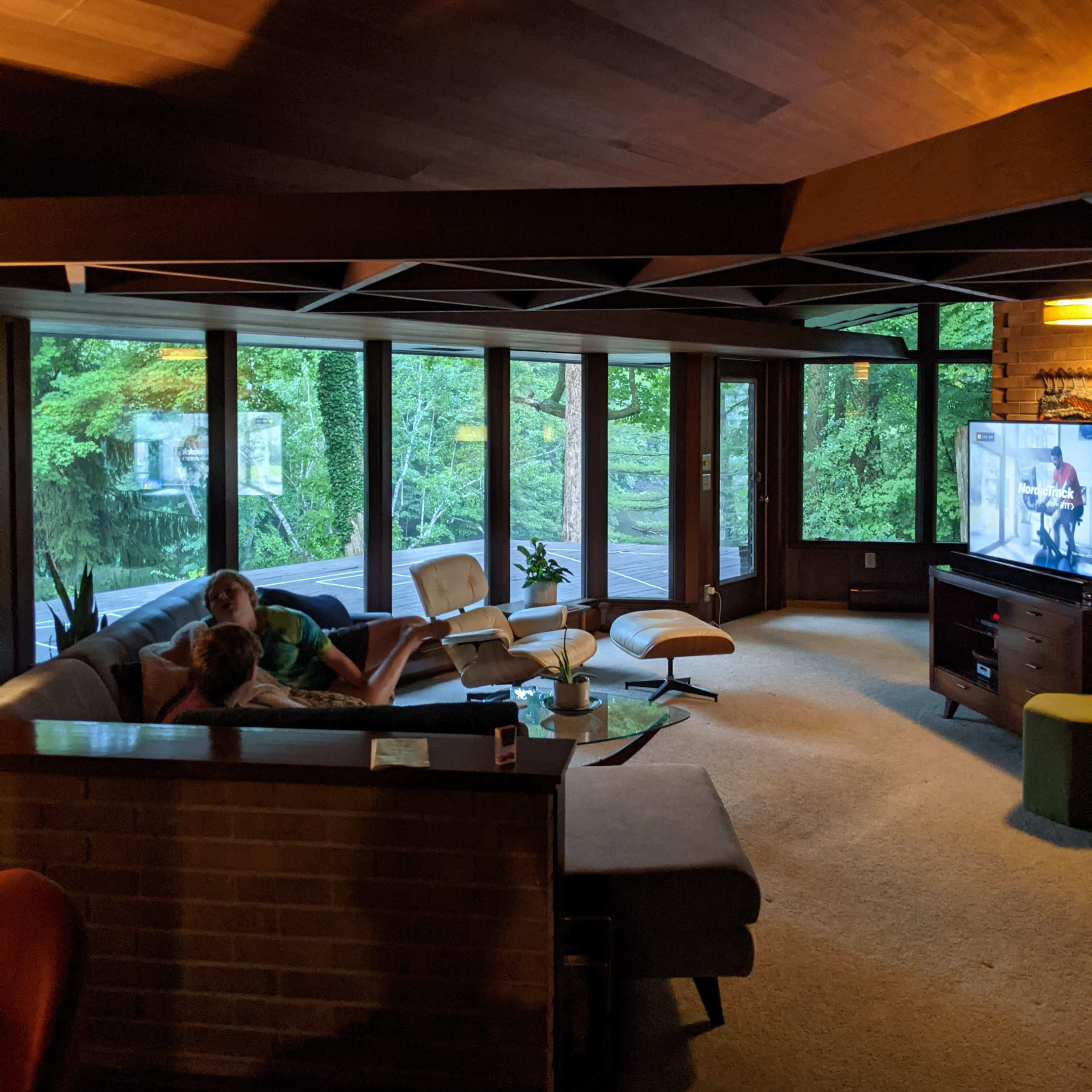 A cozy living room with large windows showcasing a view of greenery, featuring a sectional sofa, a television, and a coffee table.