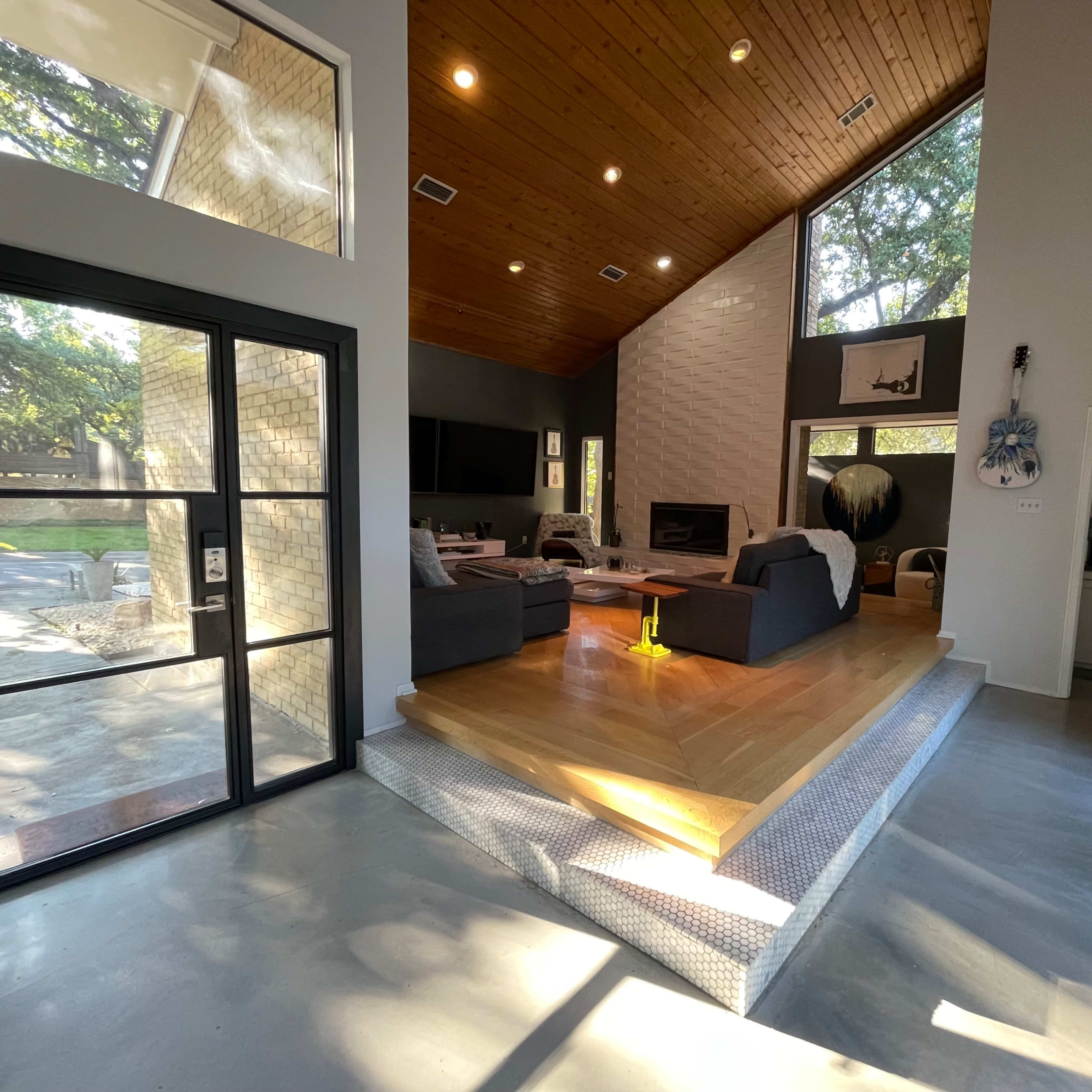 A contemporary living area with a high ceiling, a wooden beam structure, and a sunlit space featuring a fireplace and a transitional step leading to a kitchen area.
