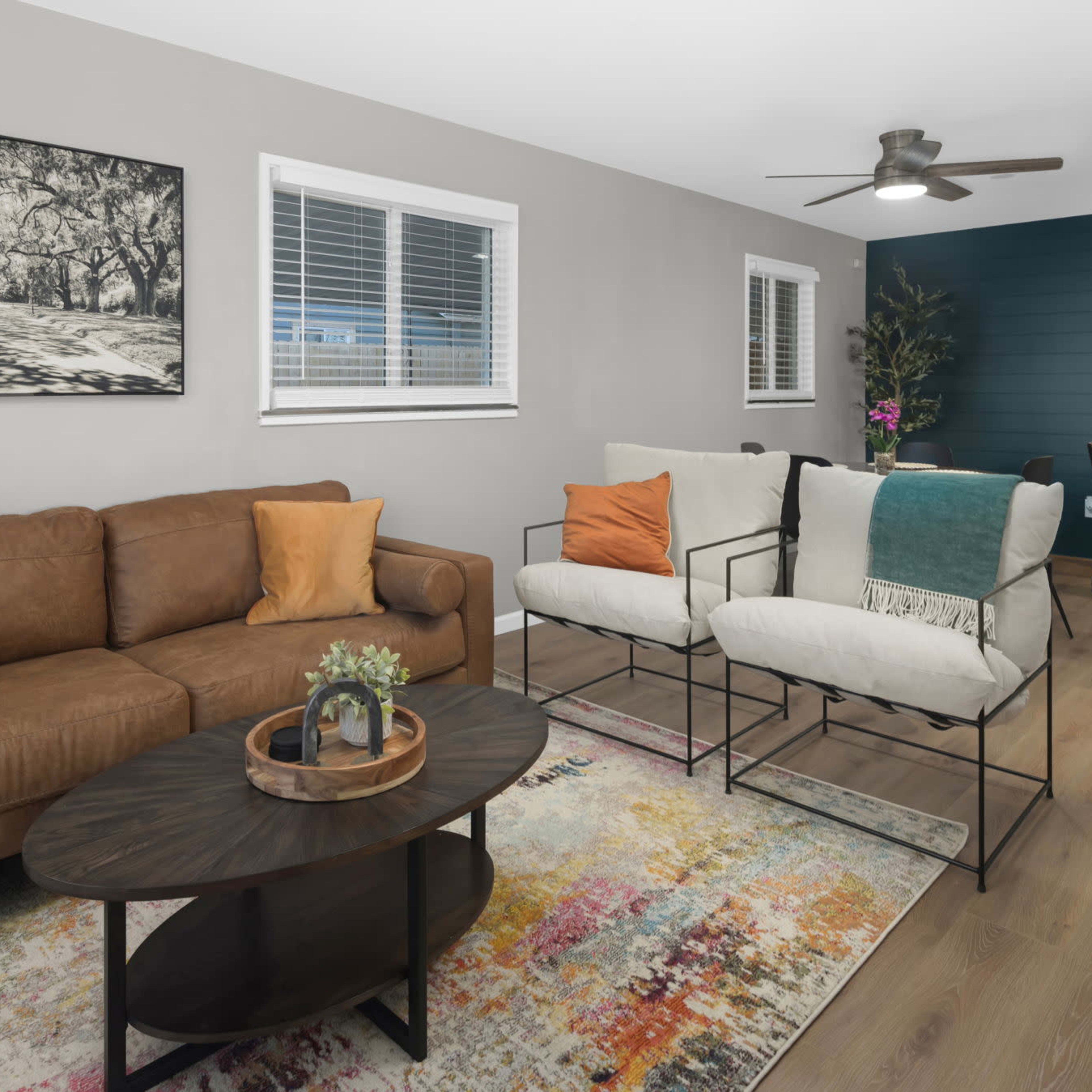 A modern living room with a brown leather sofa, two white chairs, a circular coffee table, and a colorful area rug, along with a dining area in the background.
