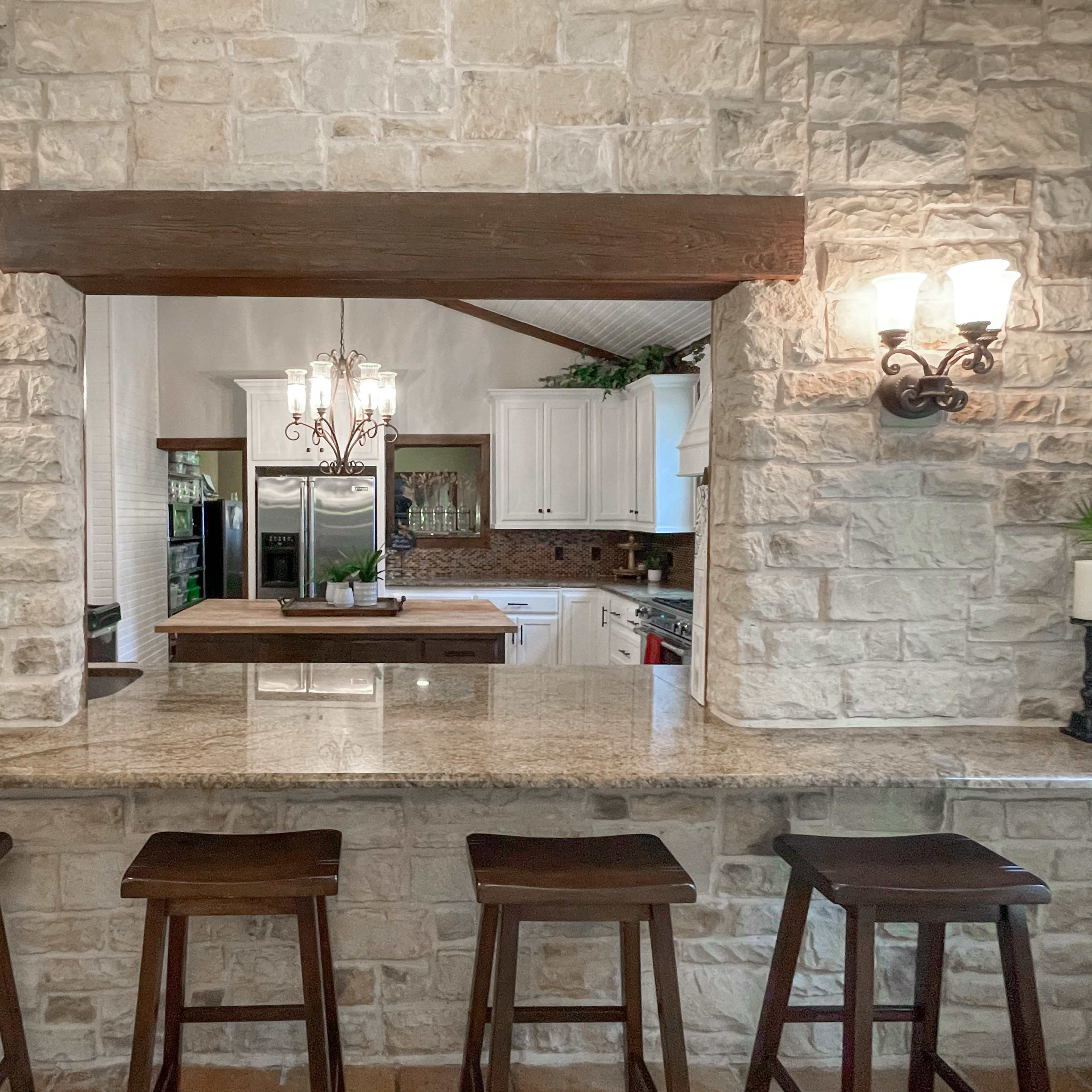 A kitchen features a stone-walled pass-through with a wooden beam, illuminated by sconces and showcasing a central island with bar stools.