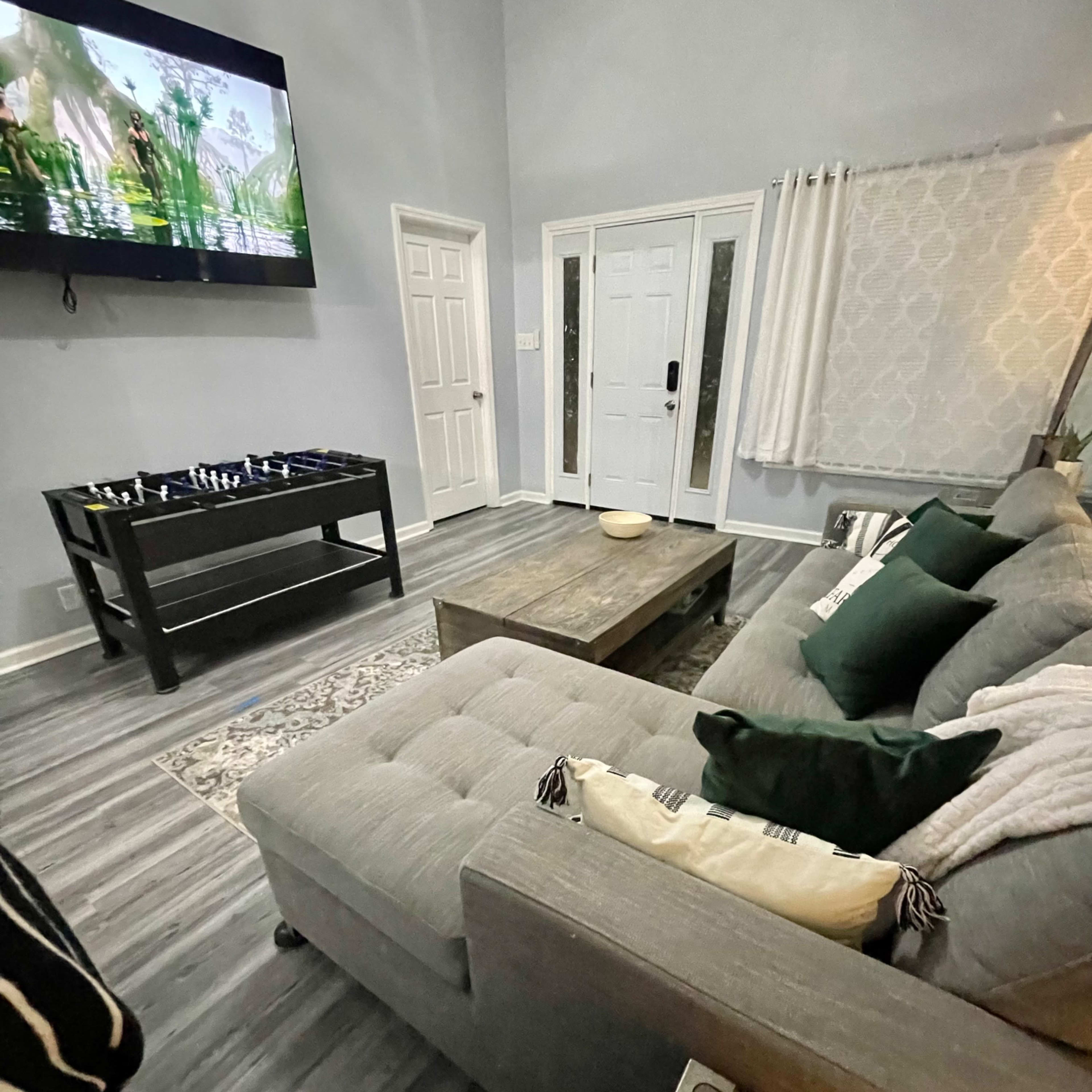 A living room featuring a grey sofa, a wooden coffee table, a television mounted on the wall, and a front door visible in the background.