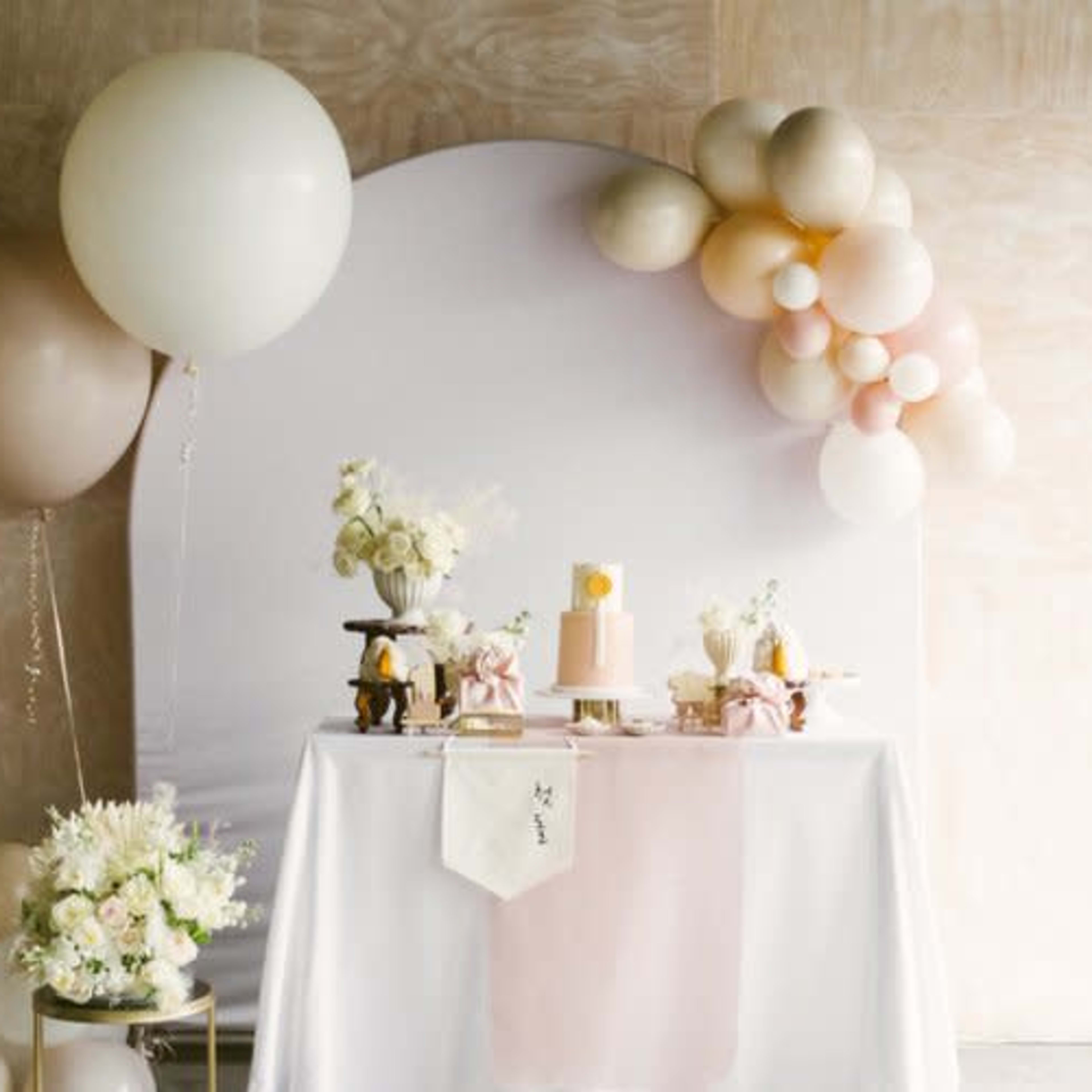 A decorative table is set up with a cake and floral arrangements, accompanied by pastel balloons and a backdrop.