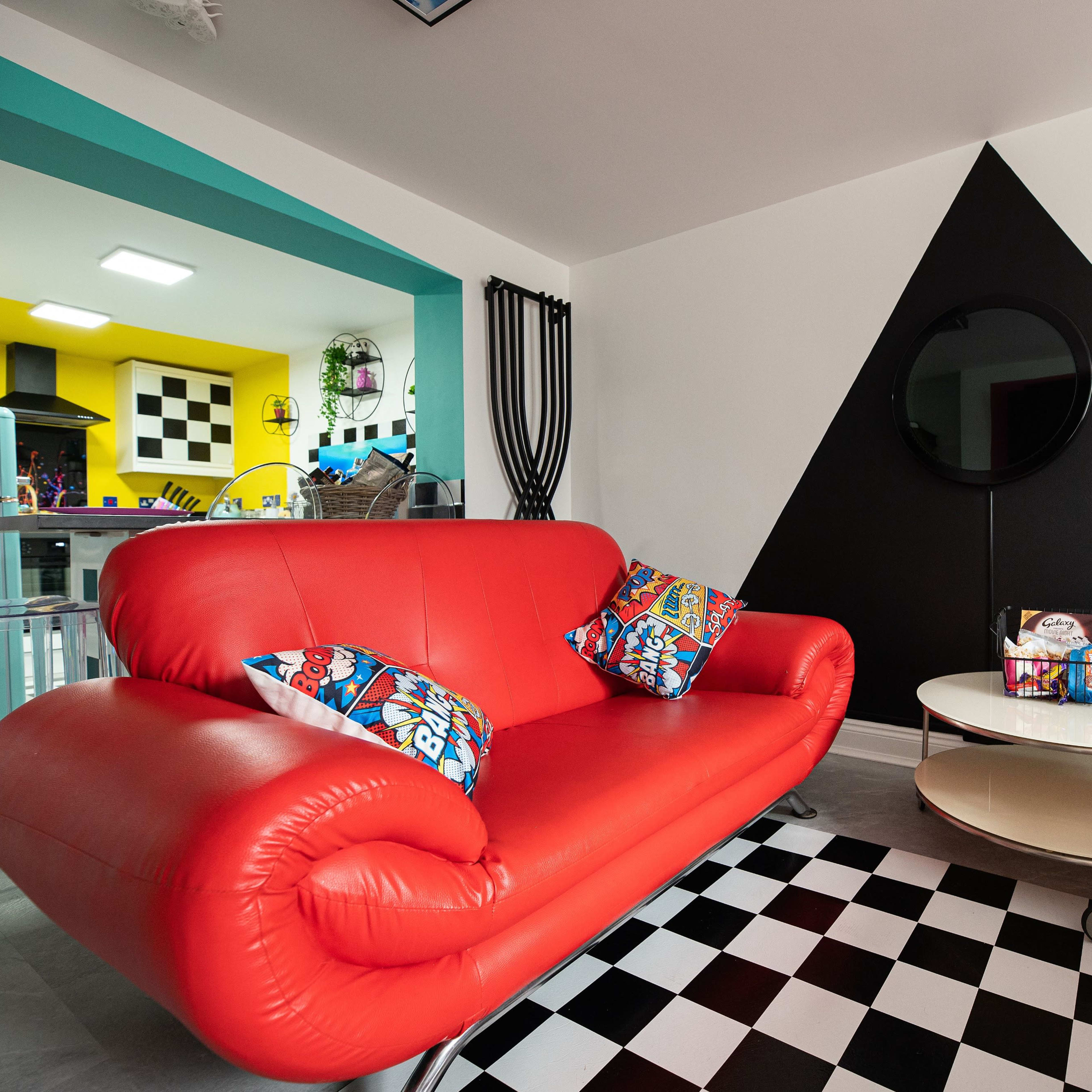 A brightly colored living space featuring red sofas with decorative pillows, a round coffee table, and a black and white checkered floor, set against a backdrop of yellow and black walls.