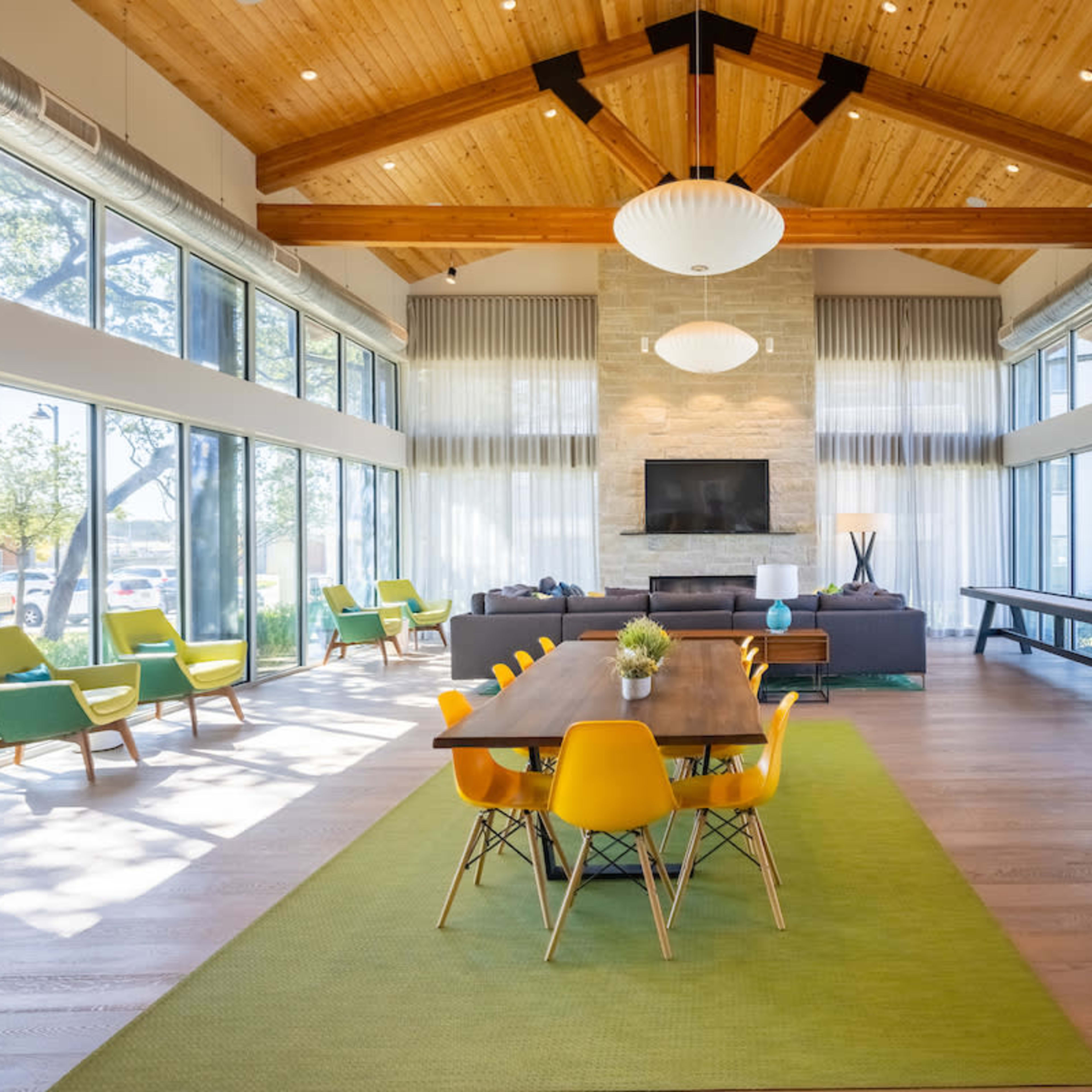 A spacious, modern lounge area with large windows, hardwood floors, a wooden ceiling, and a long dining table surrounded by colorful chairs.