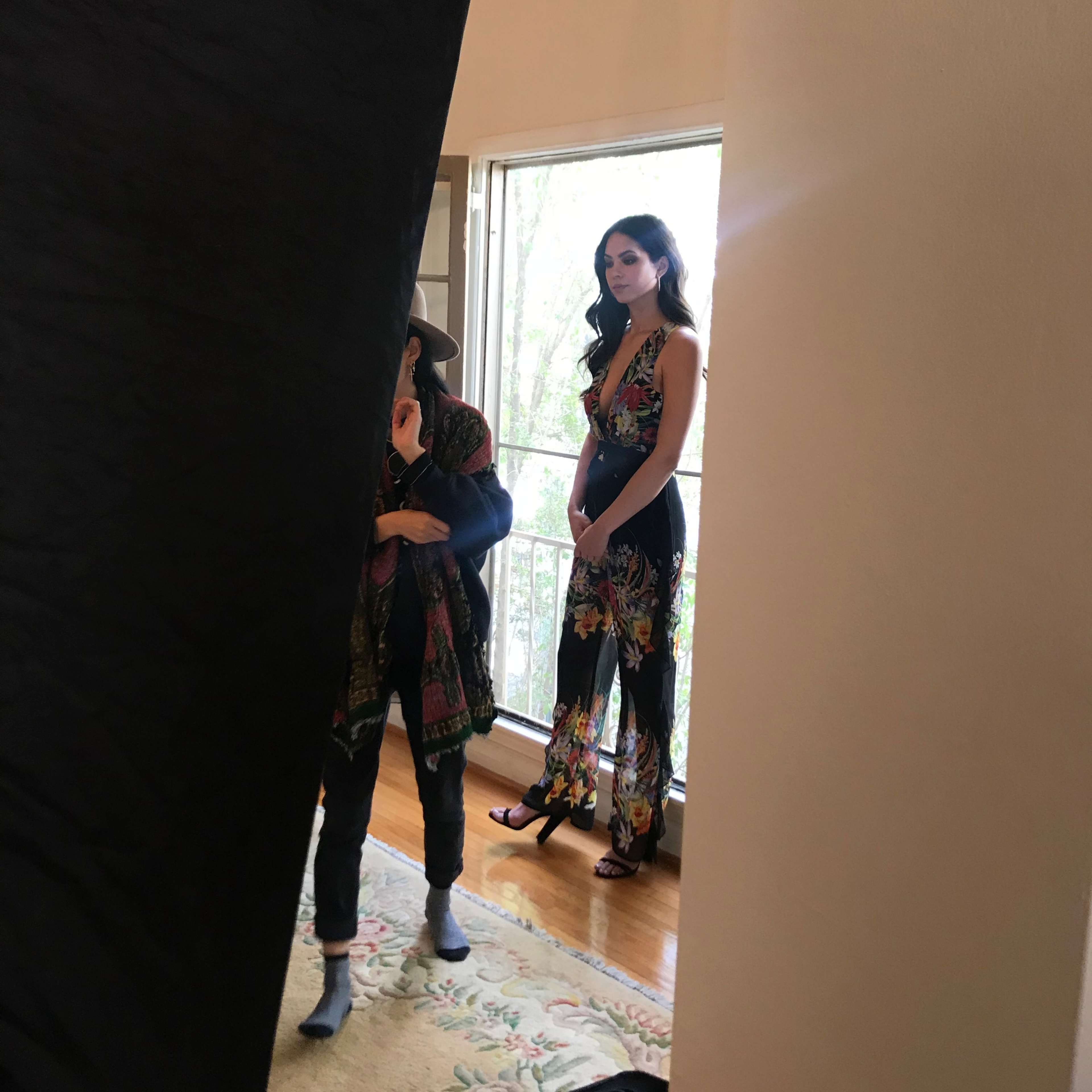 A woman in a floral outfit stands near a window while another person adjusts their attire in a room with wooden flooring and a rug.