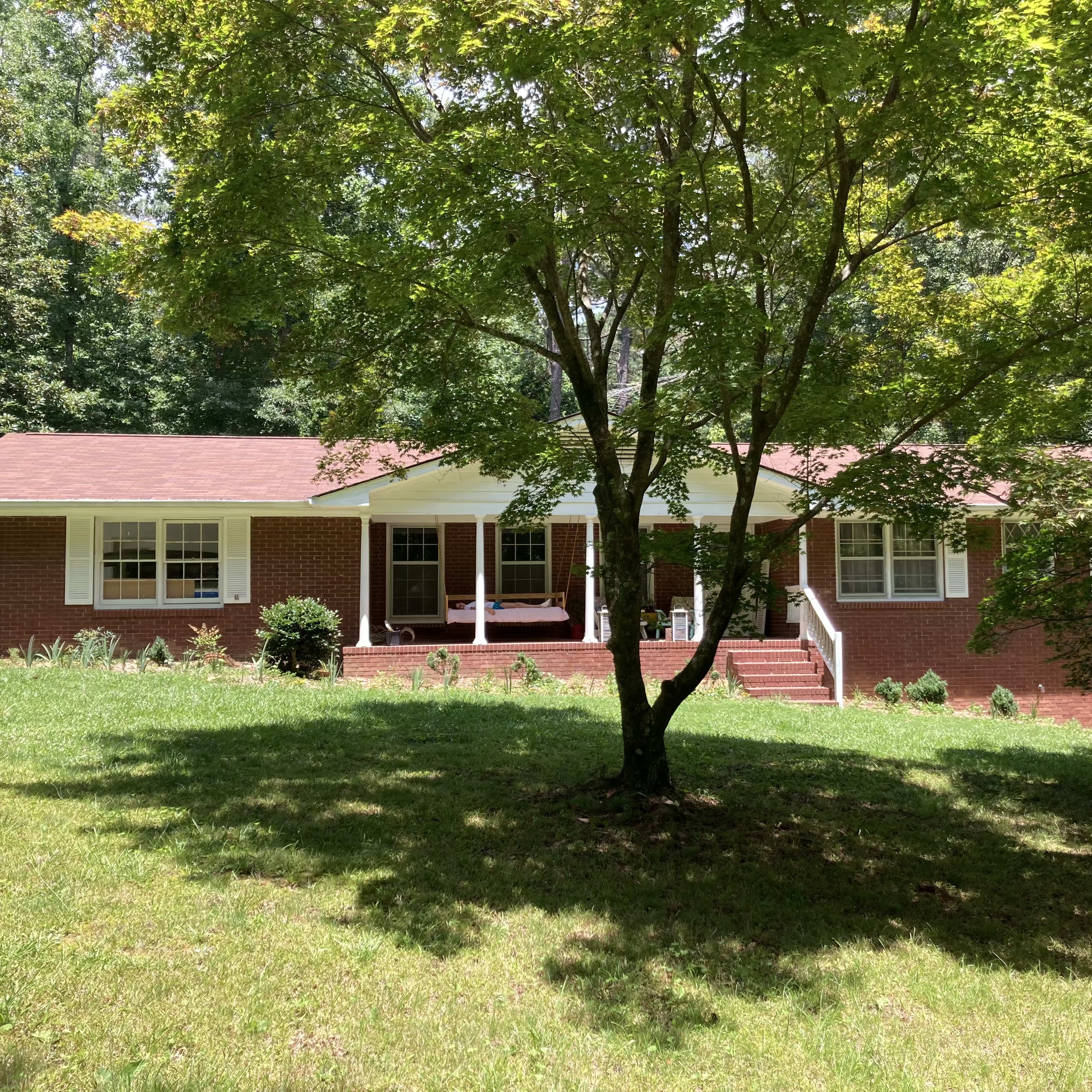 A brick house with a red roof sits on a grassy hill, surrounded by trees.
