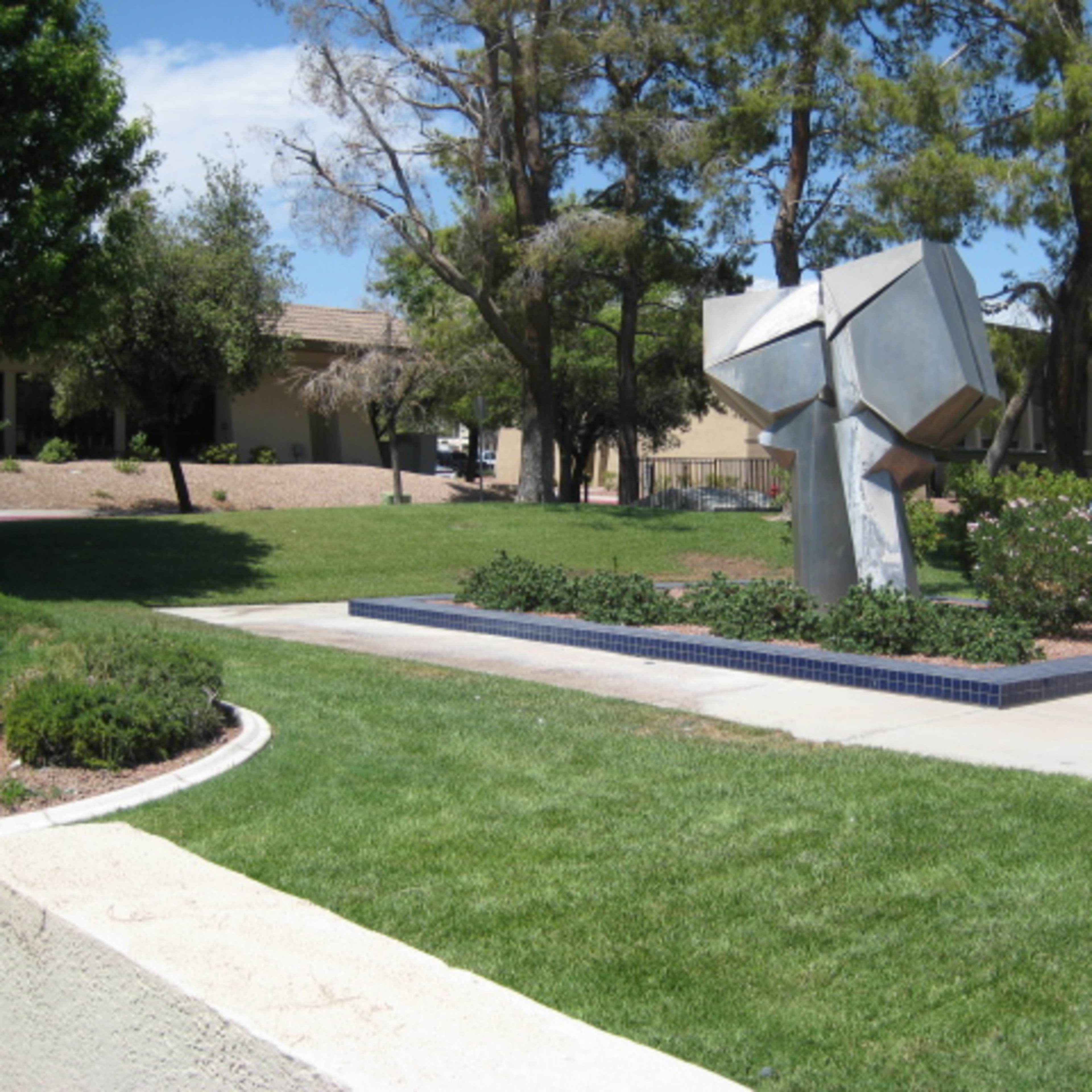 A modern metallic sculpture situated on a grassy area surrounded by bushes and trees, near a building.