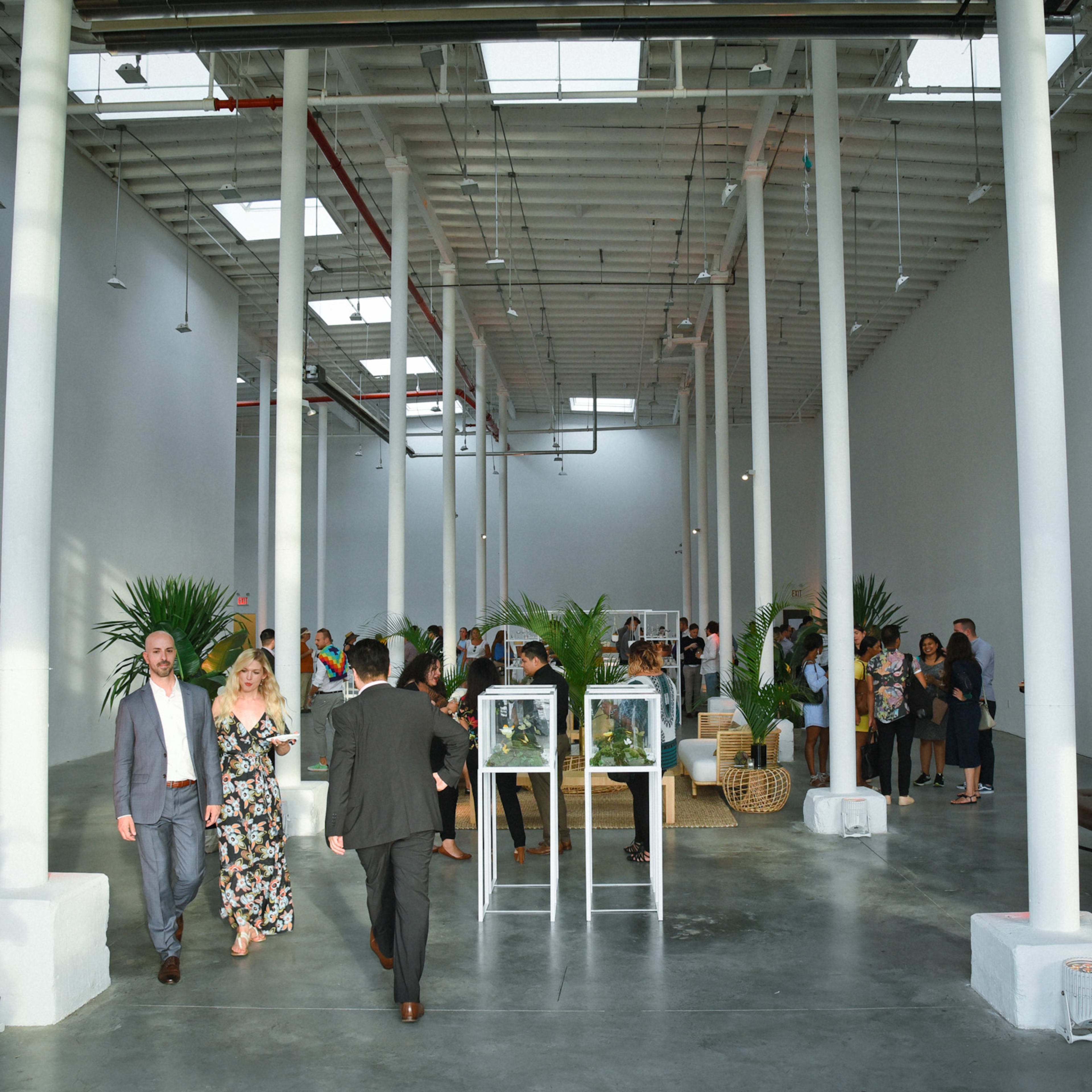 A spacious, brightly lit event venue features guests mingling, tall white columns, and modern seating arrangements.